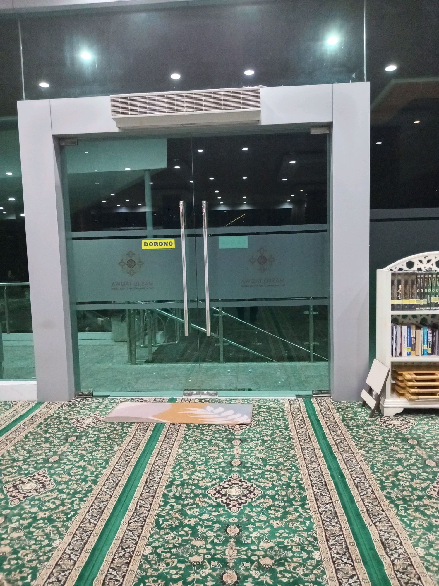 The image shows the entrance to a mosque in Yosowilangun, Indonesia. The entrance is a set of double glass doors with silver handles. A yellow sign on the door reads "DORONG," which means "push" in Indonesian. The doors are flanked by a gray frame. Inside the mosque, there is a large prayer carpet with a green and gold pattern. A small prayer mat is placed on the carpet in front of the doors. To the right of the entrance, there is a white bookshelf filled with books. The overall impression is of a clean and well-maintained religious space.