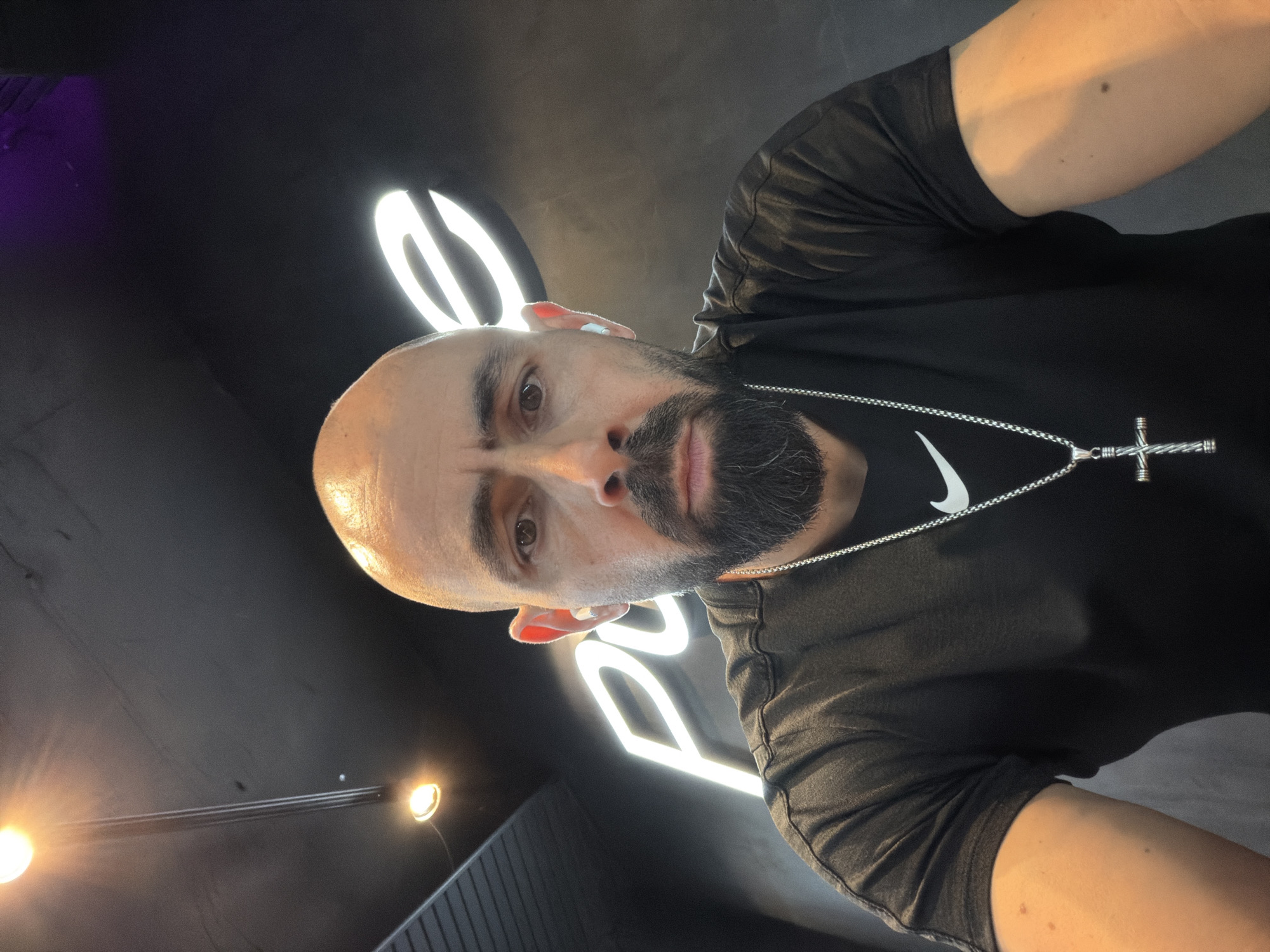A selfie of a bald man with a beard, wearing a black t-shirt and a silver necklace with a cross pendant. He is indoors, standing in front of a black wall with a white neon sign behind him. The lighting is varied, with some warm and some cool tones. The location is Plovdiv, Bulgaria.