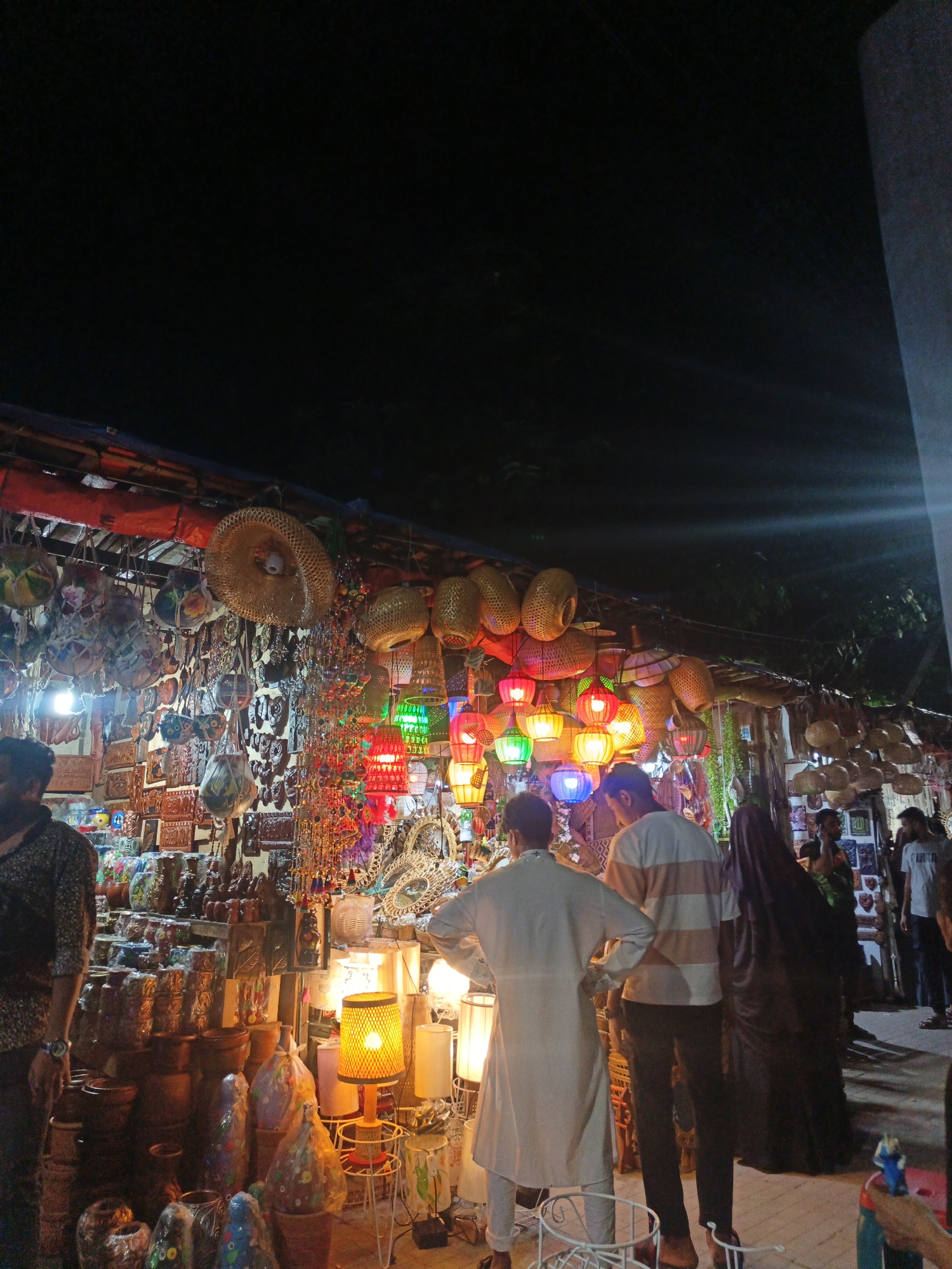 The image captures a night scene at an outdoor market stall, likely in an urban setting given the presence of streetlights and a paved ground. The stall is densely packed with a variety of handmade goods, primarily lamps and decorative items. Colorful, woven lamps in various shapes and sizes are suspended from the stall's roof, illuminated and casting a warm glow. Behind the lamps, rows of intricately carved wooden panels and decorative beads are displayed, creating a rich tapestry of textures and colors.
In the foreground, several people are browsing the merchandise. On the left, a man wearing a patterned shirt and a watch stands with his back to the camera, appearing to examine the items. To his right, two men in traditional South Asian attire, one in a long white garment and the other in a striped shirt and dark pants, are engaged in conversation or looking at the stall. Further to the right, a person wearing a dark headscarf and a long black garment is also present, as are a few other individuals in the background, some partially visible and others more clearly in frame.
The market appears to be a vibrant and bustling place, offering a wide array of artisanal crafts. The lighting, primarily from the lamps themselves and ambient street lighting, creates a somewhat dim but inviting atmosphere, highlighting the detailed craftsmanship of the goods on display. The overall impression is one of a lively night market with a focus on decorative and functional handcrafted items.