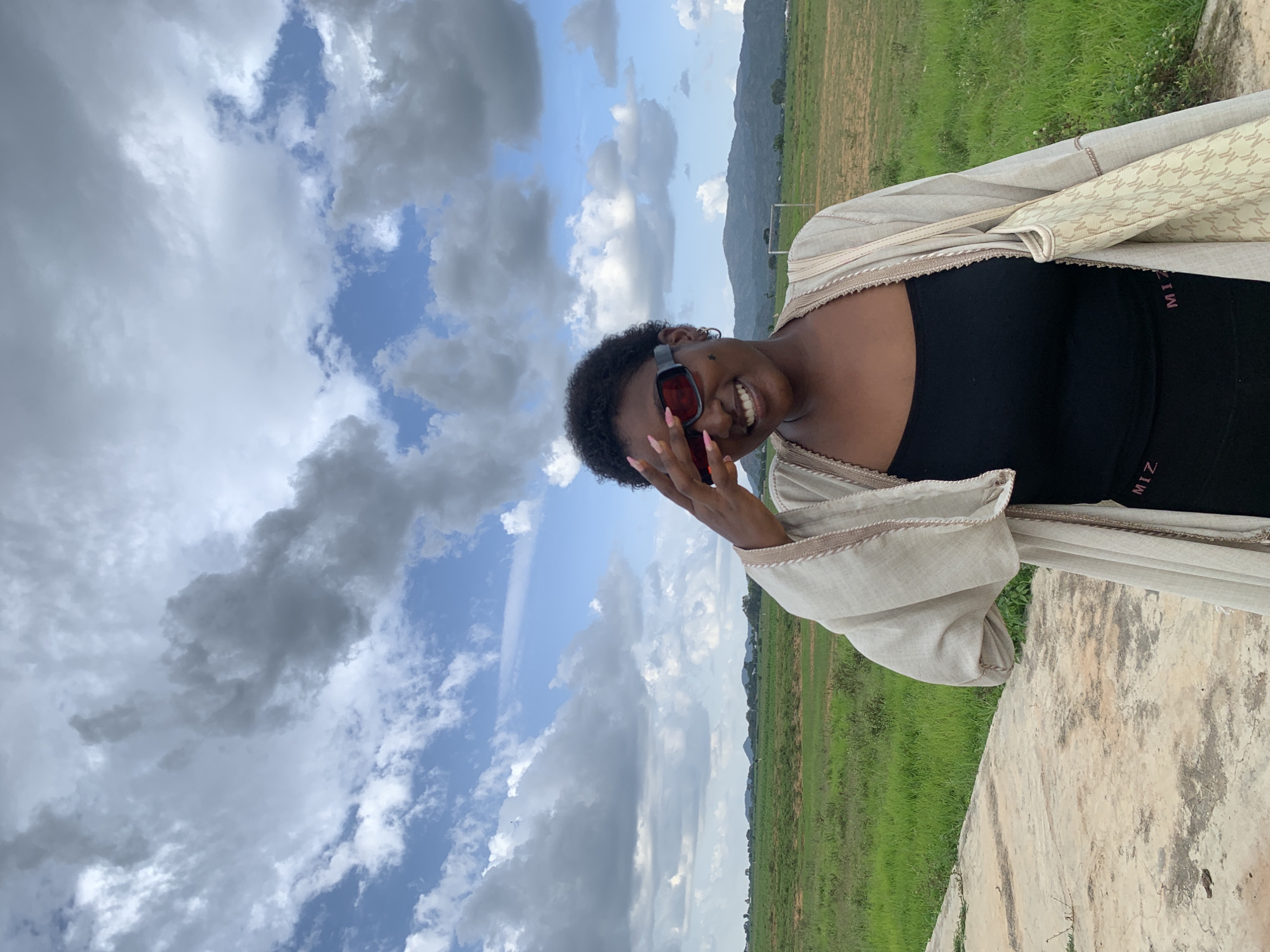A young Black woman is pictured outdoors, smiling and looking towards the camera. She is wearing a black tank top and a light-colored open cardigan with a patterned trim. Red-tinted sunglasses are on her face, and her hand is touching her forehead, partially obscuring her face. Her nails are painted a light pink.
The background features a vibrant green grassy field with some scattered white flowers, leading to a hazy mountain range in the distance. A faint outline of a soccer goal can be seen on the field. The sky is a mix of bright blue and dramatic, heavy gray clouds, suggesting a partly cloudy or potentially stormy day. The overall lighting indicates it is daytime, possibly late afternoon due to the soft light.
A concrete structure, possibly a wall or ledge, is visible in the foreground, taking up the lower right portion of the image. The photo is oriented vertically, with the subjects and background appearing sideways from a standard perspective.
Visible text on the woman's tank top reads "MIZ" in pink letters. The overall mood of the image is cheerful and relaxed, with the woman's smile conveying a sense of joy. The location context is Gwafan, Nigeria.