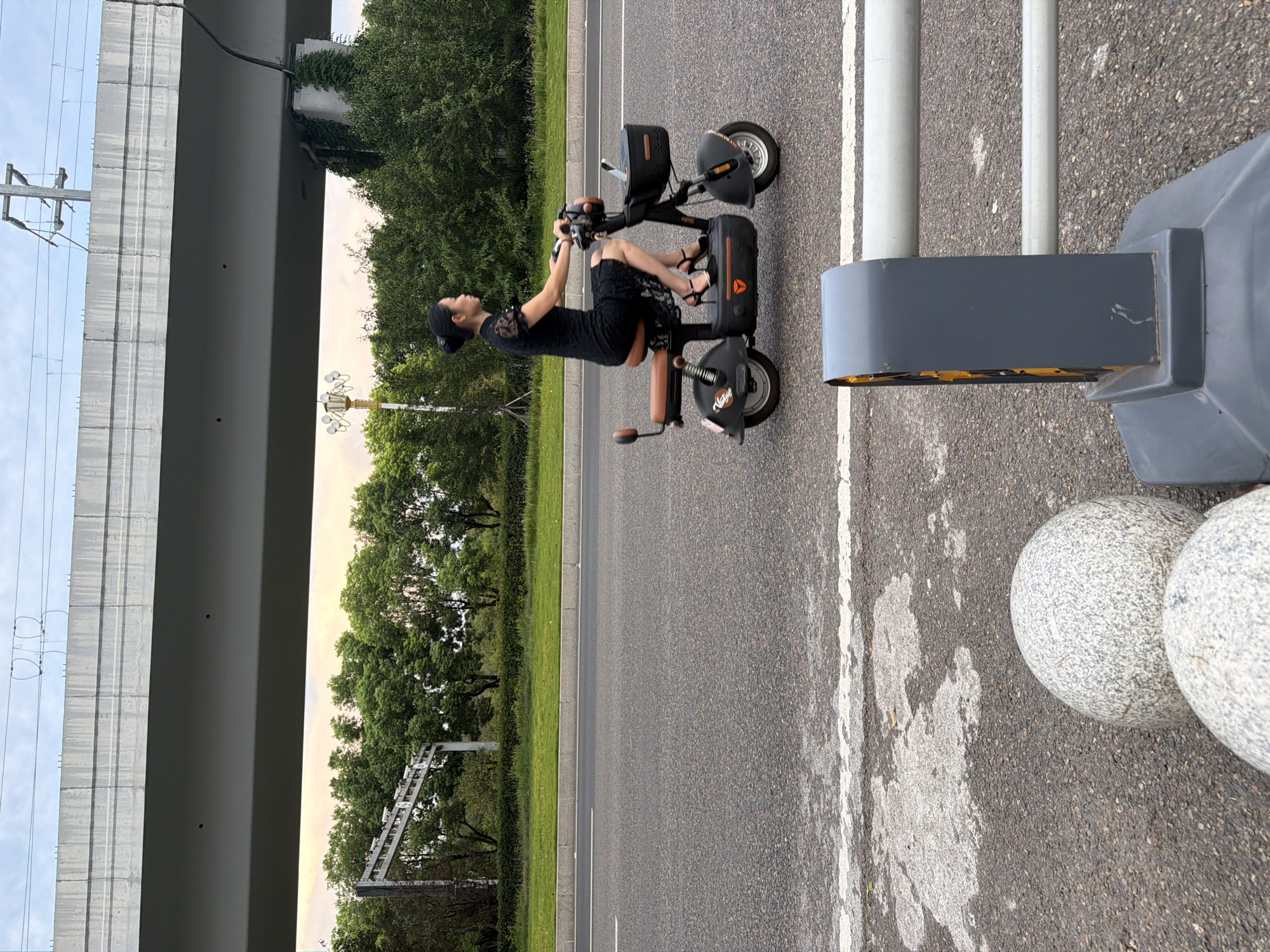A woman in a black dress rides a dark-colored electric scooter on a paved road. She is positioned in the right half of the frame, facing left, with her hands on the handlebars and her legs extended downwards. The scooter has a brown seat and handlebars, and a black basket in the front. The road surface is asphalt with white lane markings. In the background, a large concrete overpass stretches across the frame from left to right, supported by pillars. Below the overpass, there is a strip of green grass and lush trees. A vintage-style street lamp with multiple globes stands on the left side of the road. The sky is overcast with hints of yellow and orange light, suggesting either dawn or dusk. In the foreground, a grey roadside barrier with reflective yellow markings and two large, round granite spheres are visible. The scene appears to be an urban or suburban area, likely in Jiangjin, China, given the presence of the electric scooter and the style of infrastructure.