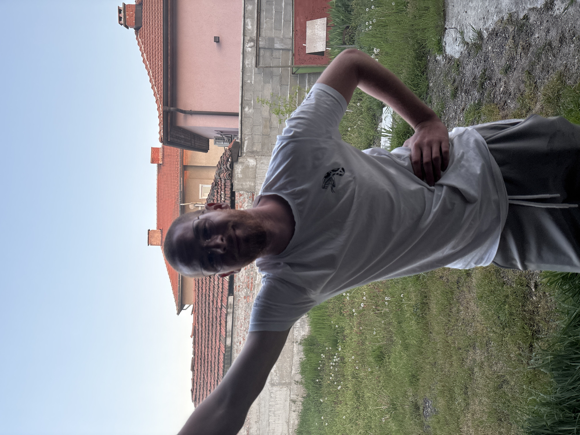 The image shows a man outdoors, seemingly in a backyard or grassy area near buildings. He is wearing a white t-shirt with a small logo on the chest and grey sweatpants. The background includes a building with a red tile roof and a pink facade. There is also a grey concrete wall. The sky is light blue. The grassy area contains patches of dandelions. The location is possibly in Chirpan, Bulgaria.