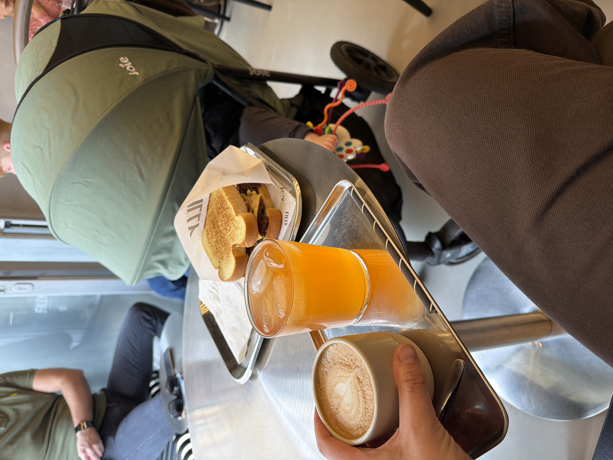 The image depicts a meal at a restaurant in Wiesbaden, Germany. The view is from a high angle, showing a metal table with a tray holding a sandwich, a glass of iced orange juice, and a cup of coffee with latte art. A person is holding the cup of coffee. A baby stroller with a green canopy is positioned next to the table. A baby can be seen reaching for a toy attached to the stroller. The background includes a window with a partial view of the outside. Another person is seen wearing a green shirt and black pants, sitting at the table. Someone else is wearing brown pants.