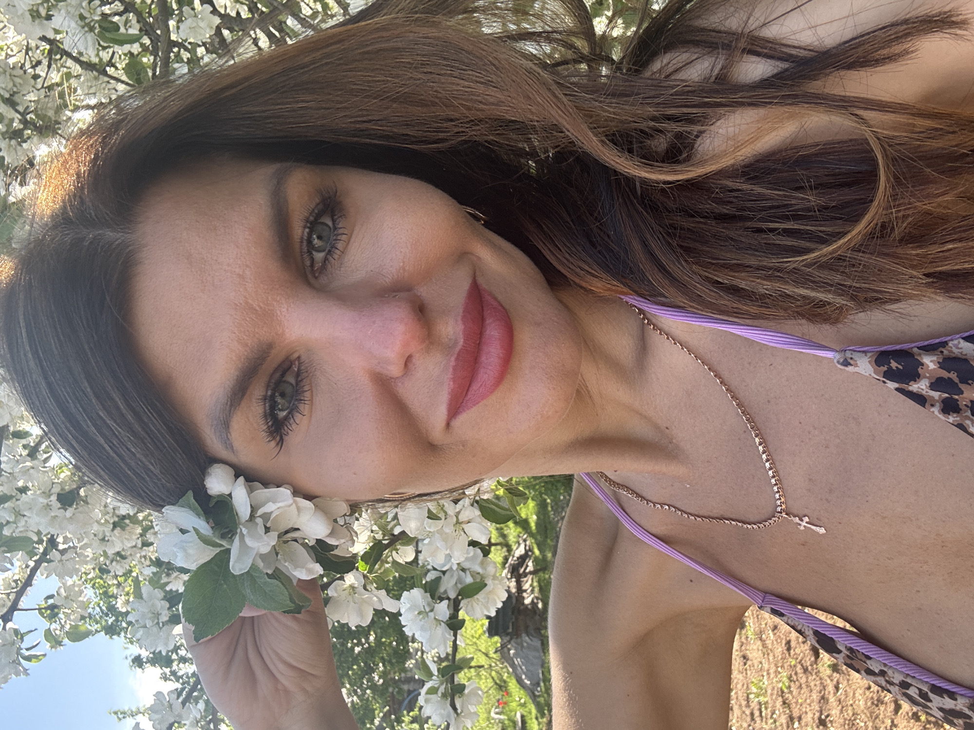 A woman with brown hair and green eyes poses for a selfie in a garden setting. She is holding a branch of white flowering blossoms near her face. She is wearing a purple and brown patterned top, and a delicate gold necklace with a cross pendant. The background features more flowering trees and greenery. The location is outdoors, presumably in a garden or park. There is no specific information present in the image that identifies the location as the city of Dubovaya, Russia.