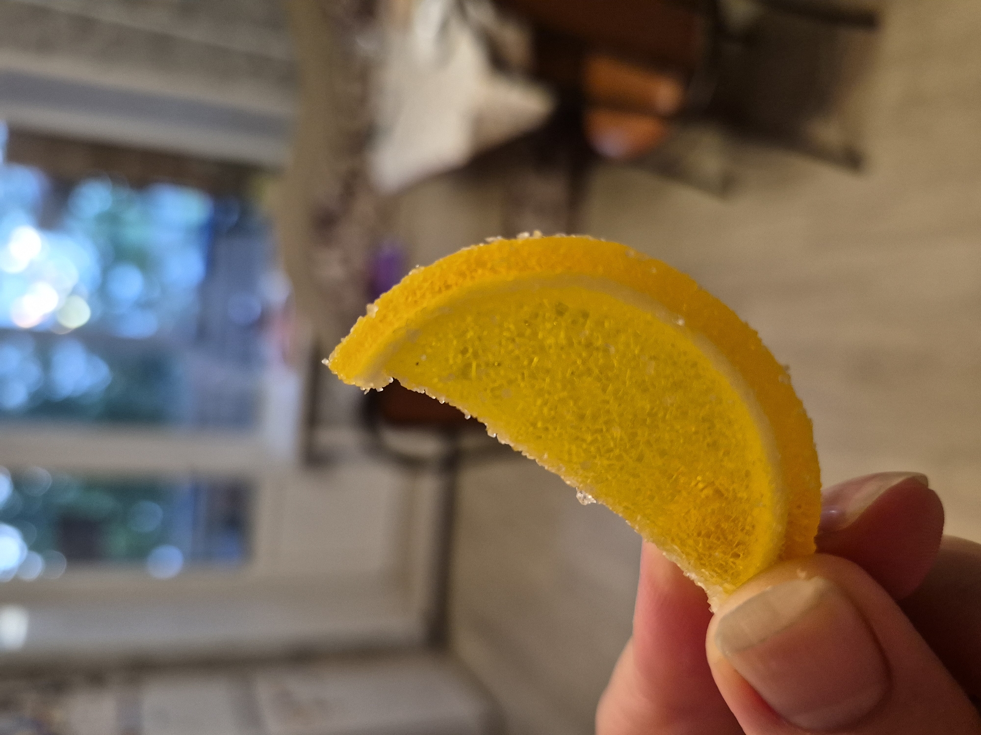 A close-up shot shows a person holding a crescent-shaped, candied orange peel slice between their fingers. The peel is a vibrant yellow-orange color and is dusted with sugar crystals, giving it a slightly textured appearance. The surface of the peel is translucent, and some small air bubbles are visible within its sugary matrix. The hand holding the peel has manicured fingernails, and the skin appears to be of light to medium tone.
The background is out of focus, suggesting a shallow depth of field. To the left, a blurred window reveals a bright, outdoor scene with dappled light and greenery, indicative of daytime. The lighting suggests indoor ambient light mixed with daylight coming through the window. The overall mood appears casual and relaxed, with no specific event or emotion being conveyed. The setting appears to be domestic, possibly a kitchen or living area, given the blurred elements in the background that could be furniture or household items. No text is visible in the image. The location is specified as Nyagan, Russia.
