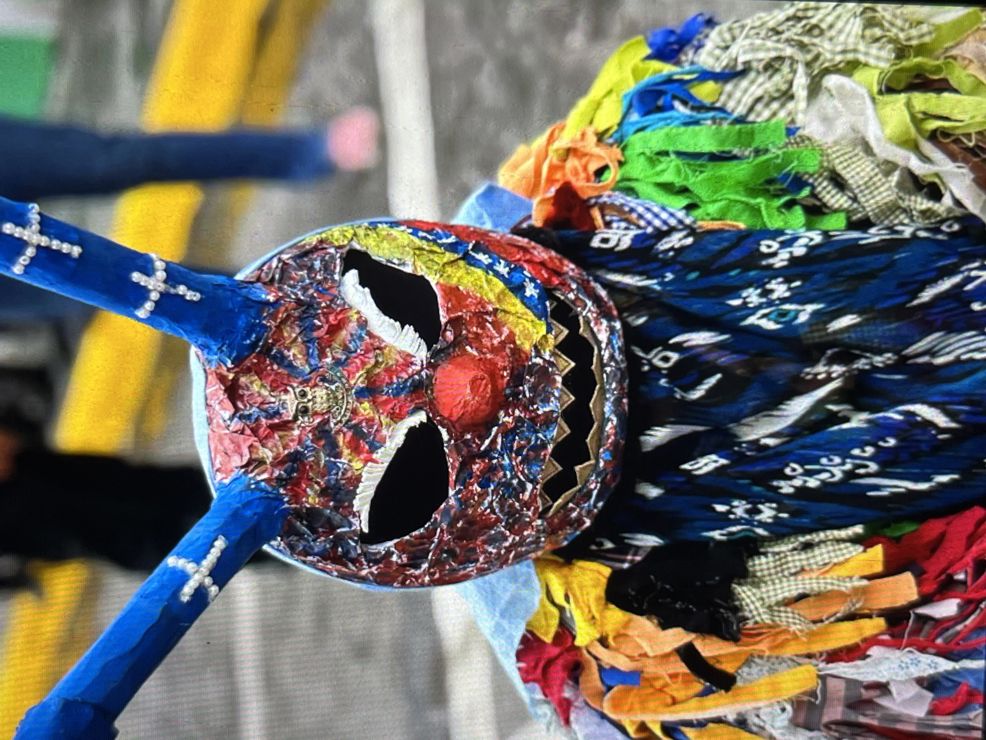 The image shows a close-up of a person wearing a colorful, elaborate mask and costume, likely during a festival or parade. The mask itself is brightly colored with red, blue, yellow, and other hues, featuring large, dark eye holes and a prominent red nose. It also has two long, blue, horn-like protrusions adorned with white cross-like decorations. The costume consists of a patchwork of various fabrics and colors, with many strips of cloth hanging from the shoulders and chest, creating a vibrant and textured appearance. The background suggests an outdoor urban setting, with blurred figures and yellow and grey lines possibly indicating a street or sidewalk. The scene appears to be in Tinaquillo, Venezuela, as indicated by the location reference.