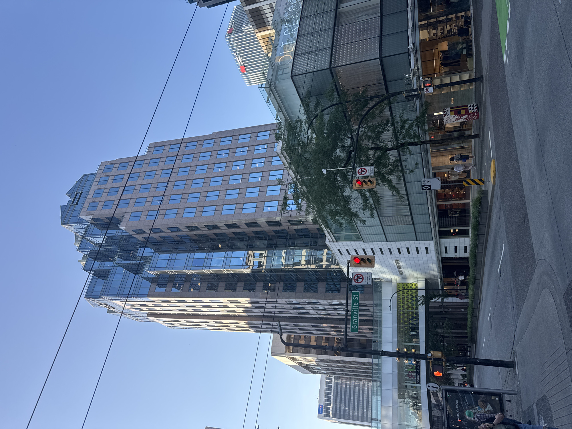 This image depicts an urban street scene in Vancouver, Canada, specifically at the intersection of Granville Street and a cross-street. The foreground shows a street with a crosswalk and sidewalks. A traffic light is visible, indicating a red light. Overhead, power lines traverse the sky.
Dominating the scene are modern, multi-story buildings. The primary building is a tall, contemporary office tower with a facade of glass and a tan-colored material. Its upper section features a stepped, angular design. Adjacent to this tower is a lower, glass-fronted building that appears to house retail establishments, with the name "HOLT RENFREW" illuminated on its facade. Further in the background, other office buildings are visible, including one with the "NATIONAL BANK" logo.
Several people are present. On the sidewalk in the mid-ground, two individuals are walking away from the camera. On the right side of the street, more people are visible near the entrance of a store, appearing to browse or enter. In the lower left corner, a person is partially visible, walking towards the camera.
The environment is urban, with clear blue skies overhead. Trees line the street, adding some greenery to the concrete and glass landscape. The overall impression is of a bustling downtown commercial district.