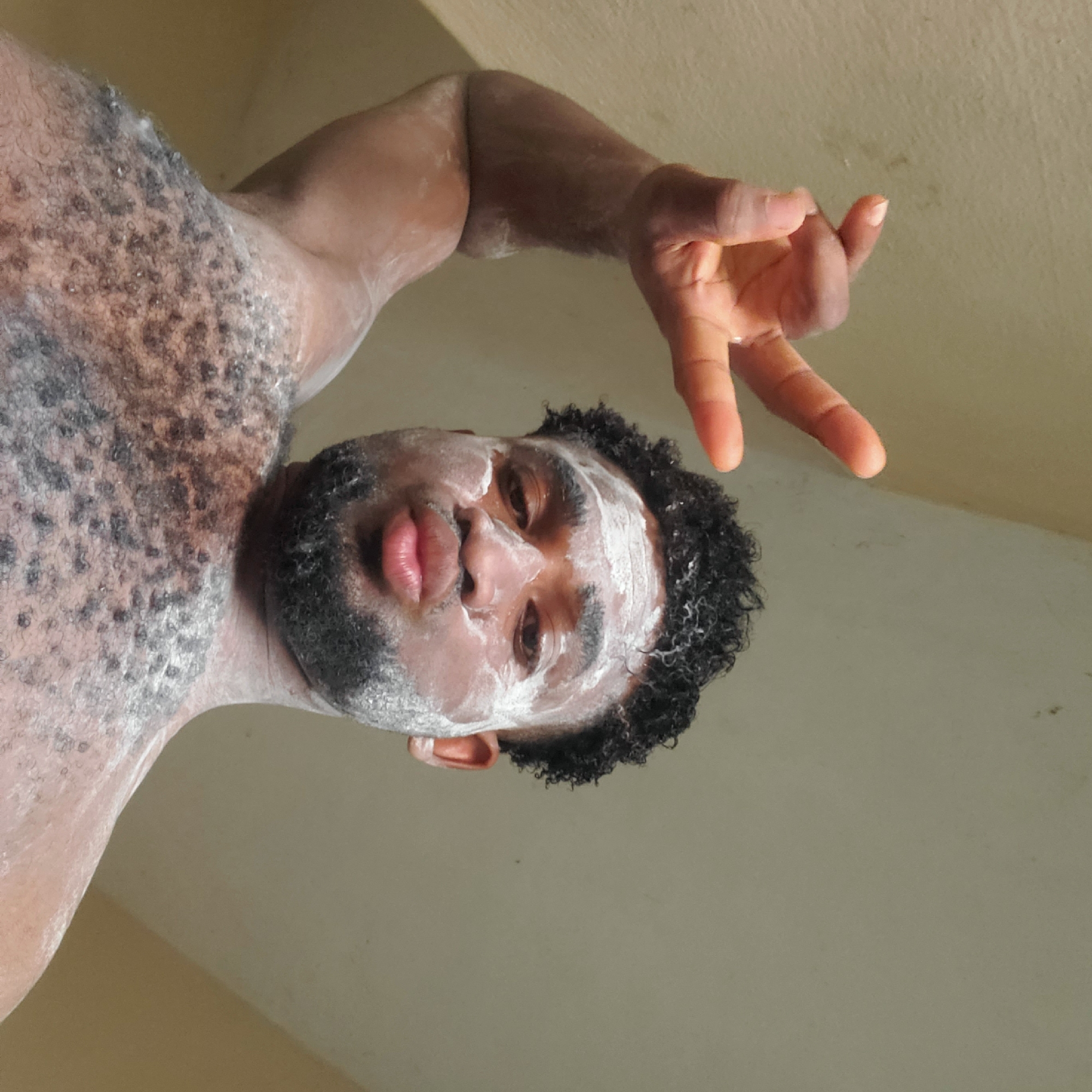 A dark-skinned man with short curly black hair is positioned in the lower center of the frame, angled upwards towards the camera. His face is partially covered with a white substance, possibly cream or powder, which also appears on his chest and arms. He has a beard and mustache. His lips are pursed. His right hand is raised with the index and middle fingers extended. The background appears to be a light beige wall or ceiling. There are no other visible people or objects. The location in Obosi, Nigeria is not apparent from the image.