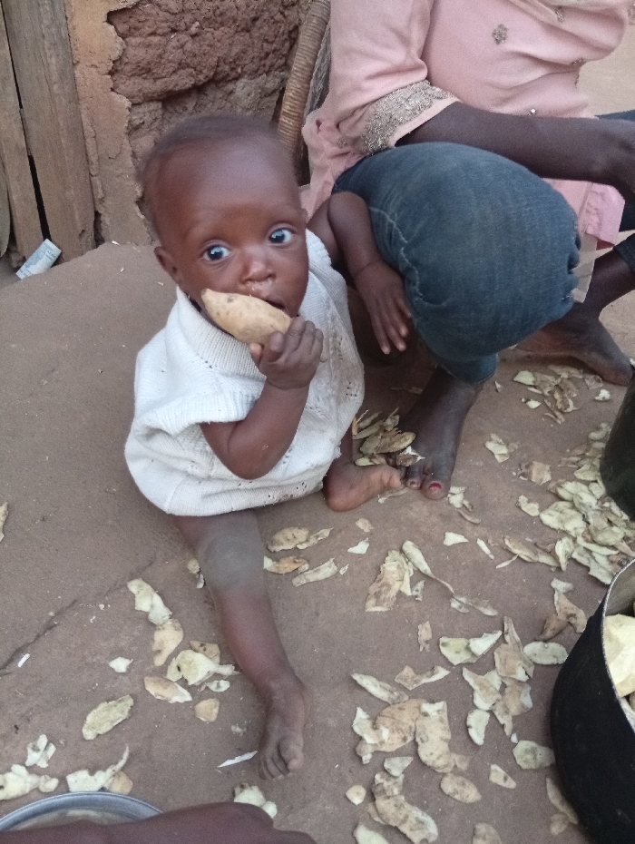An infant of African descent, wearing a light-colored knitted top, sits on reddish-brown dirt ground in Unguwan Rimi, Nigeria. The infant holds a light-brown, elongated root vegetable to its mouth and looks directly at the camera with wide eyes. Surrounding the infant are scattered light-colored vegetable peels. A partially visible adult, also of African descent, is seated to the infant's right, wearing blue denim trousers and a pink-hued embroidered garment, with bare feet visible. A dark-colored pot containing additional root vegetables is on the ground to the right, and a metallic bowl is partially visible in the bottom left. The background consists of a rustic wall constructed from reddish-brown earth, with wooden planks on the left side.