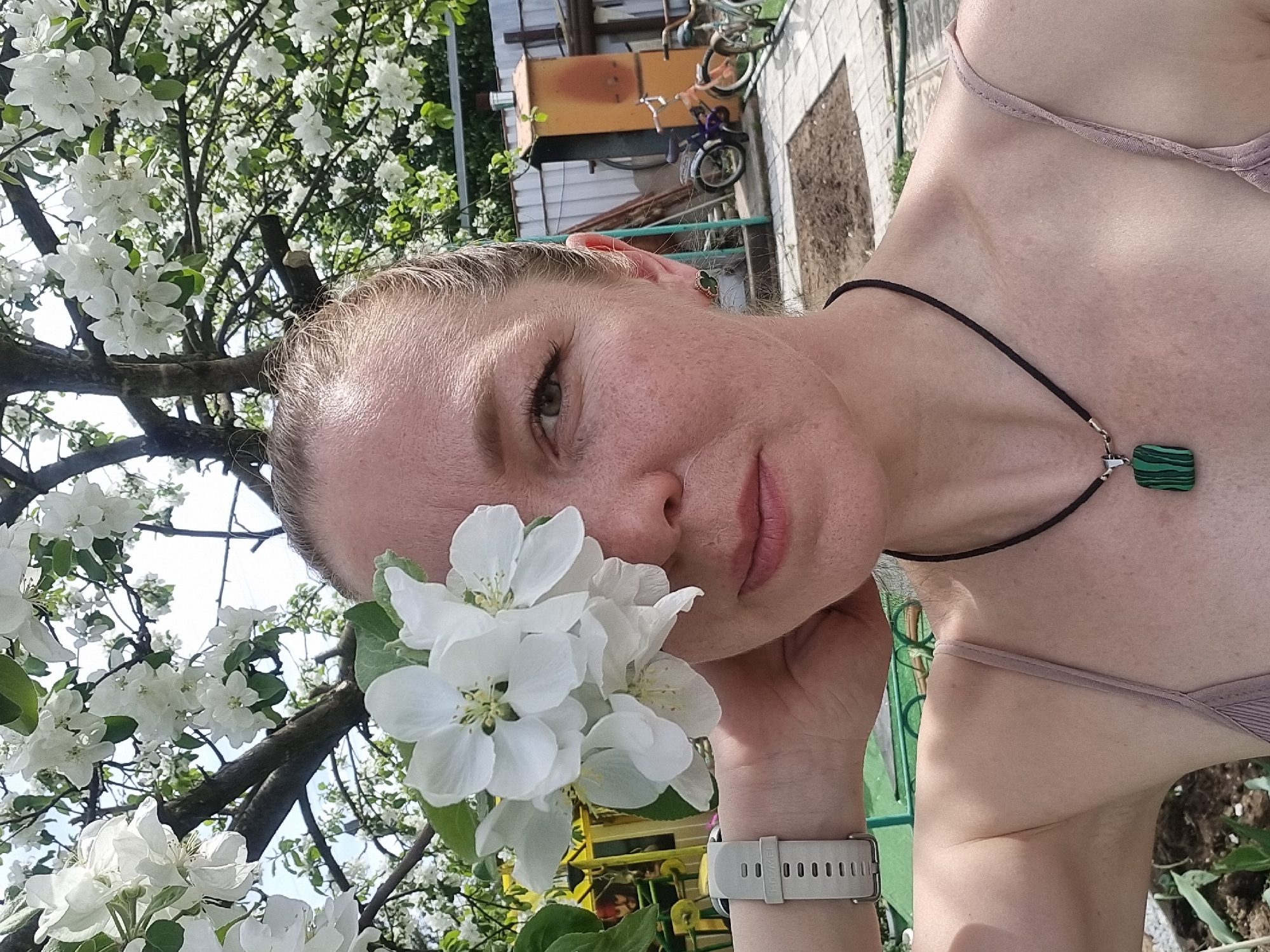 A fair-skinned woman is holding white flowers in front of her face. She is wearing a neutral-colored tank top and a black cord necklace with a green and black rectangular pendant. She also has a watch with a light-colored band on her wrist. Behind her, there is a tree in full bloom with white flowers. In the background, there is a building, a bicycle, and a patch of bare earth. The picture was taken in Chaikovsky, Russia.