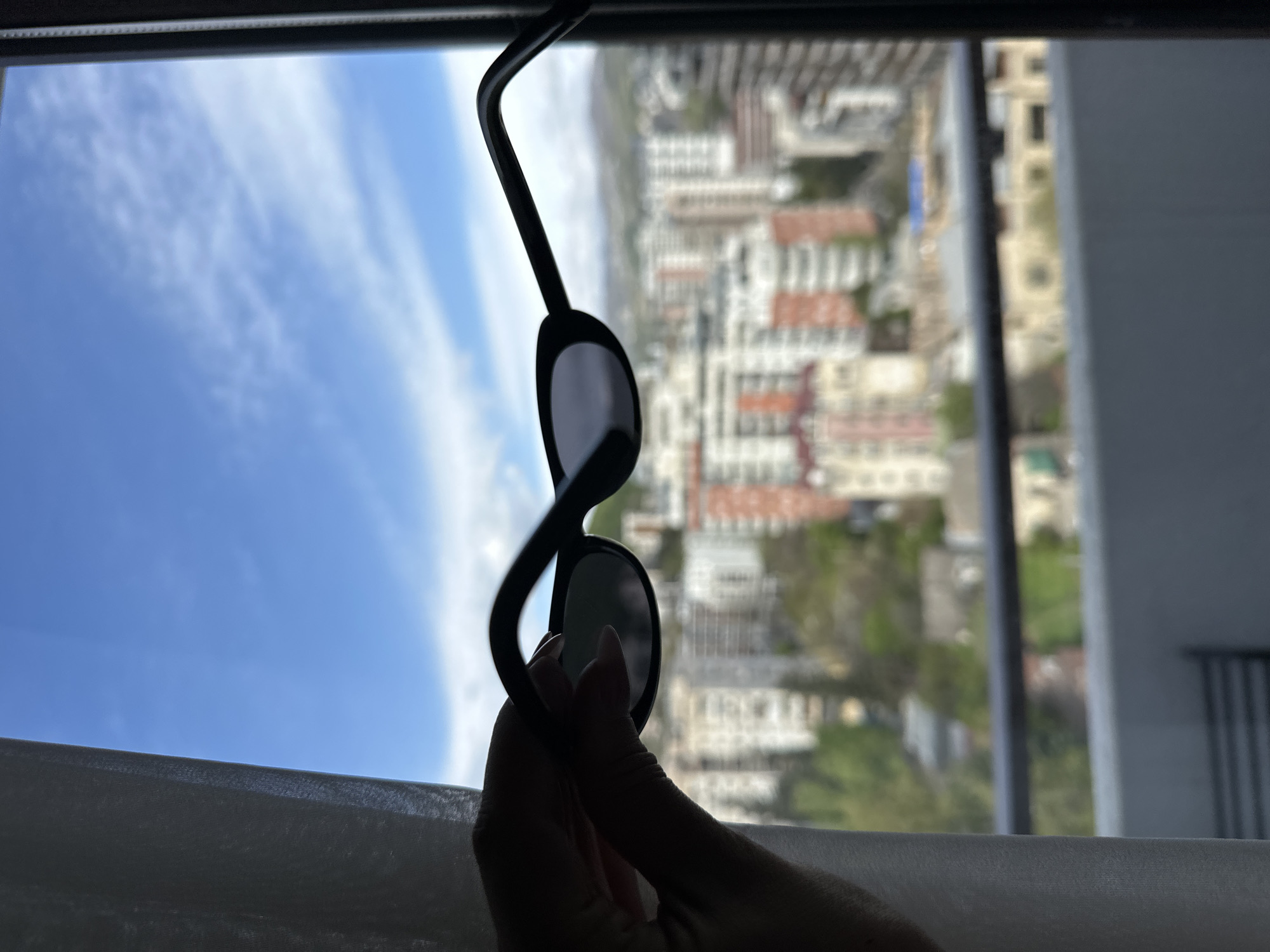 A person's hand is holding a pair of dark sunglasses in front of a window, with a view of Tbilisi, Georgia, visible through the lenses and around the frame. The background shows a cityscape with buildings of varying heights and colors, including red-tiled roofs. The sky above the buildings is blue with scattered white clouds. The curtain in the foreground is white, partially obscuring the view.