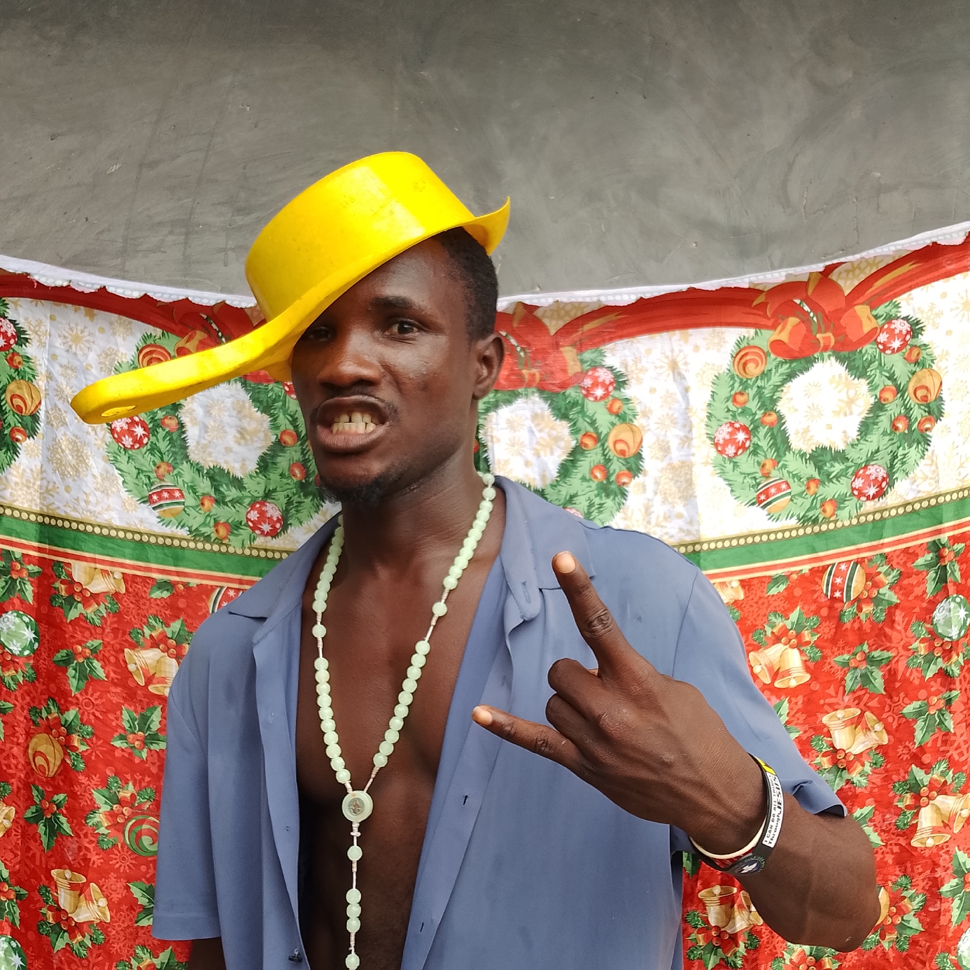 A medium shot captures a dark-skinned man from Ado, Nigeria, wearing a blue collared shirt unbuttoned at the neck, revealing a light green beaded necklace with a circular pendant. He sports a bright yellow bowl-shaped hat tilted precariously on his head. The man's mouth is open, displaying his teeth, and he is making a "rock on" hand gesture with his right hand. His left hand is raised, with his index finger and thumb forming a circle. Behind him, a backdrop features a festive red and green Christmas-themed pattern with wreaths, bells, and holly. The ground is not visible in the frame.
