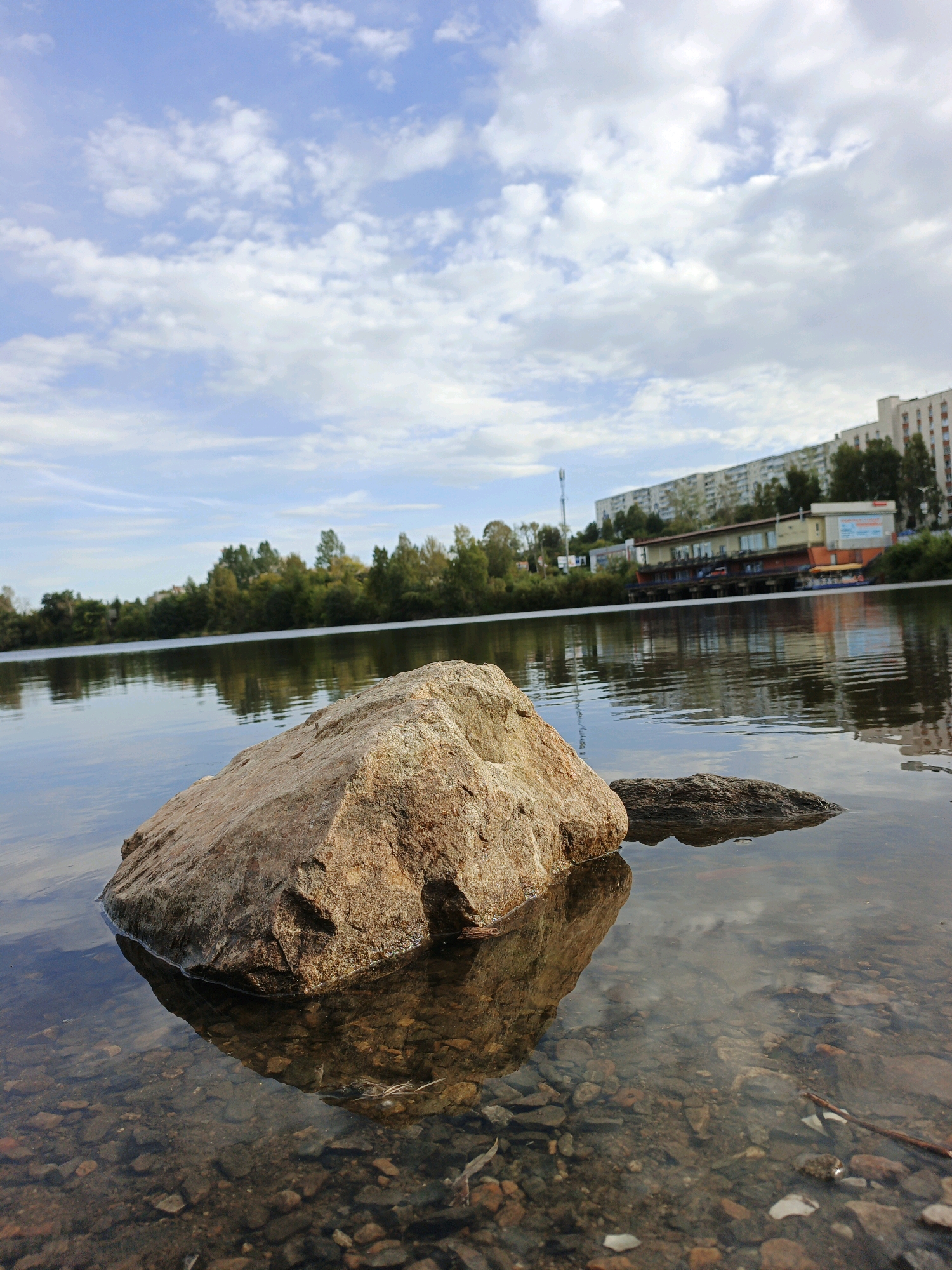 A body of water, likely a river or lake, dominates the foreground of the image. In the shallow water near the camera, a large, rough-hewn, tan-colored boulder rests on the rocky riverbed. The water is clear enough to see the pebbles and smaller stones beneath the surface. Another smaller, darker rock is visible to the right of the large boulder, partially submerged.
Beyond the immediate foreground, the water stretches back, reflecting the sky and the distant shore. The far bank of the water body is lined with a dense band of trees, exhibiting various shades of green and some hints of autumn yellow.
In the background, a long, multi-story building complex, characteristic of Soviet-era architecture, stretches horizontally across the scene. Some parts of this building appear to be residential, while others seem to be commercial or recreational, with visible awnings and signage. A tall communication tower stands to the left of the buildings. The sky above is a light blue, with scattered white, cumulus clouds. The overall impression is a view from the waterfront looking towards an urban landscape, suggesting the presence of Zlatoust within or adjacent to this natural setting. There are no people visible in the image.