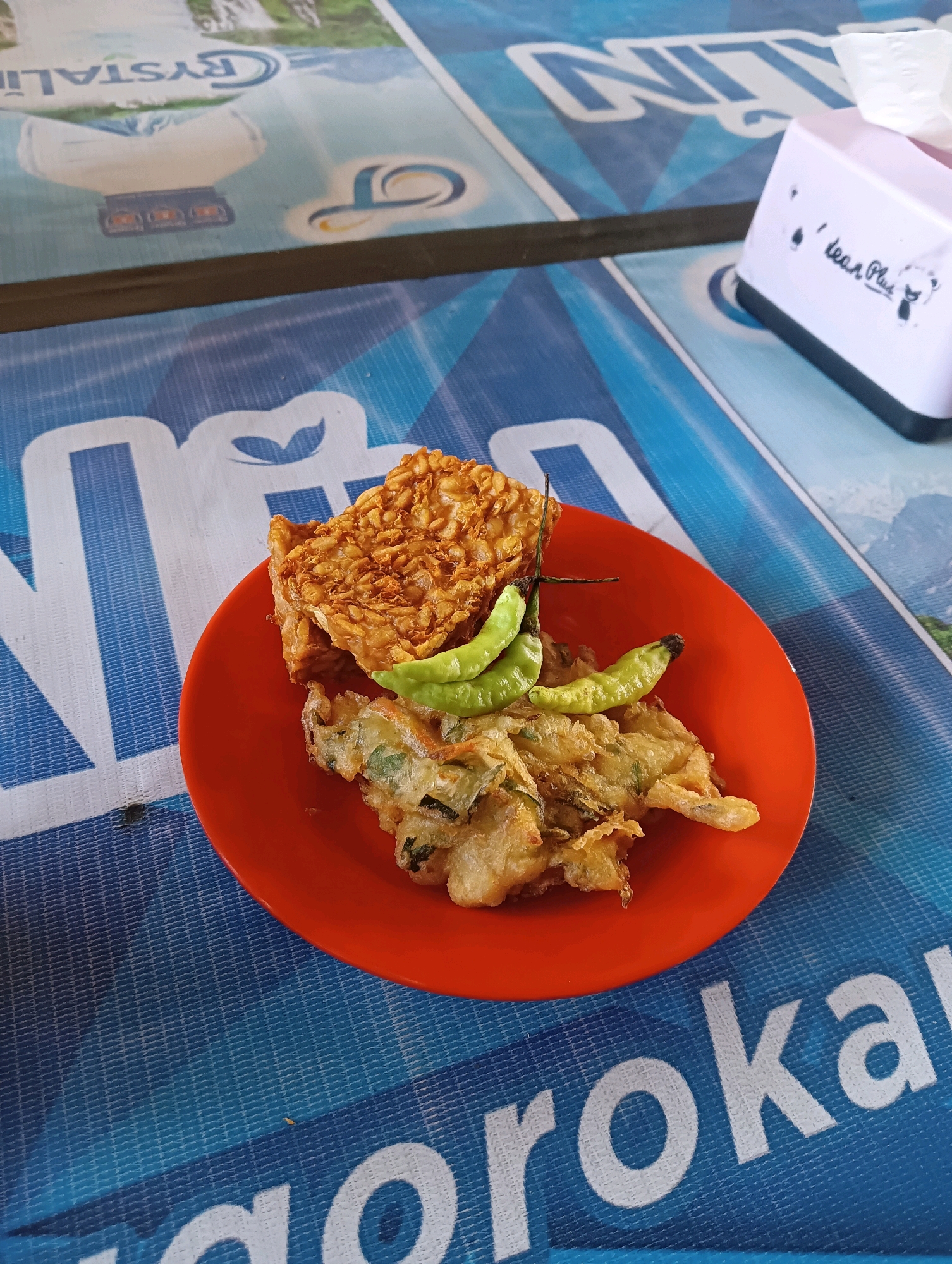 A close-up, overhead shot shows a bright orange plate holding a serving of Indonesian food. The plate sits on a blue and white patterned table. The food consists of several pieces of fried tempeh, a common Indonesian soy product, and some fried fritters containing vegetables. Two small, light green chili peppers are placed on top of the tempeh. In the background, a white tissue box with some writing and a small drawing is visible on the right side of the table. The table itself has large, white text printed on it, partially visible, which appears to be part of a logo or branding. The overall scene suggests a casual dining setting, likely in a street food stall or a local eatery within Kecamatan Pati, Indonesia, given the context.