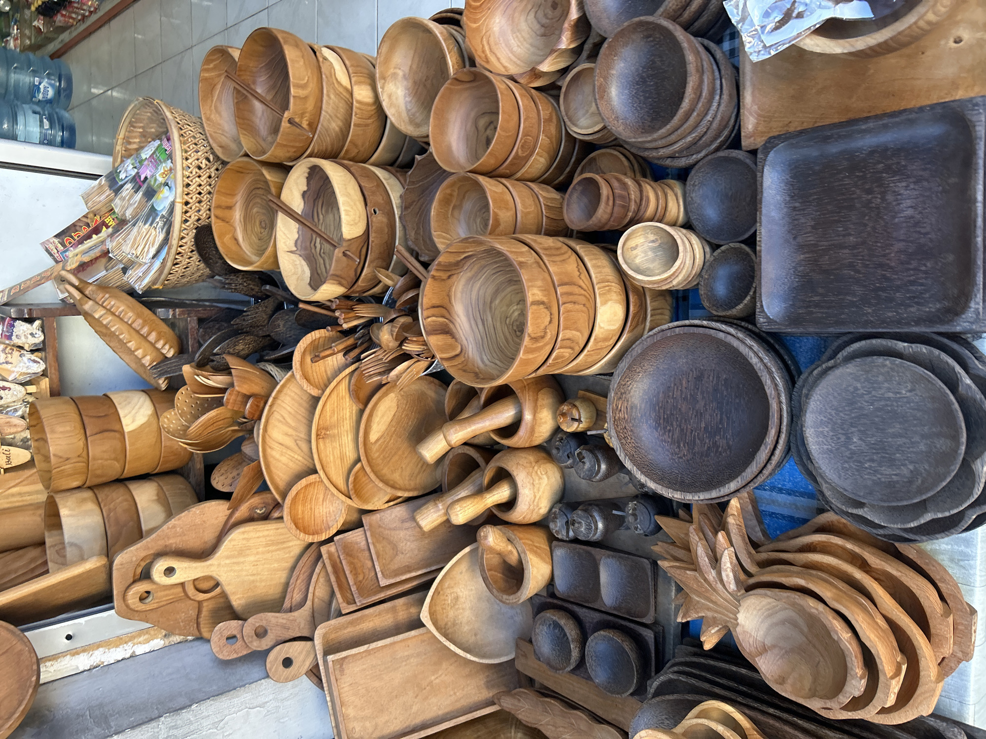 This image depicts a street-side stall in the city of Ubud District, Indonesia, showcasing a variety of handcrafted wooden kitchenware. The stall is filled with an assortment of bowls, platters, cutting boards, and mortars and pestles, all made from different types of wood with varying finishes. In the background, stacks of bottled water and other items suggest it's part of a larger marketplace or shop. The scene is well-lit, likely by natural daylight, highlighting the natural grains and textures of the wood. There are no people visible in the immediate foreground or mid-ground.