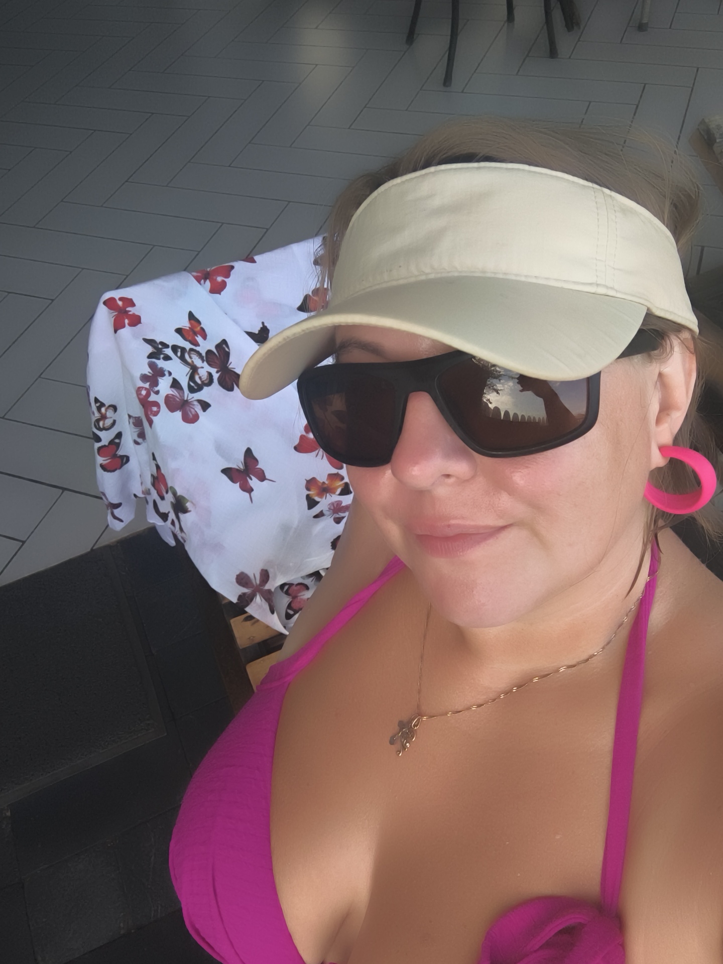 A medium shot shows a woman wearing a pink bikini top, a beige visor, and large dark sunglasses. She has bright pink hoop earrings. A thin gold chain with a small charm hangs around her neck. The background includes a white cloth with a butterfly print and a tiled floor. The overall scene is bright and suggests an outdoor setting, possibly a poolside or patio area. There is no identifiable information about the city of Phu Thuy Ward, Vietnam in this image.