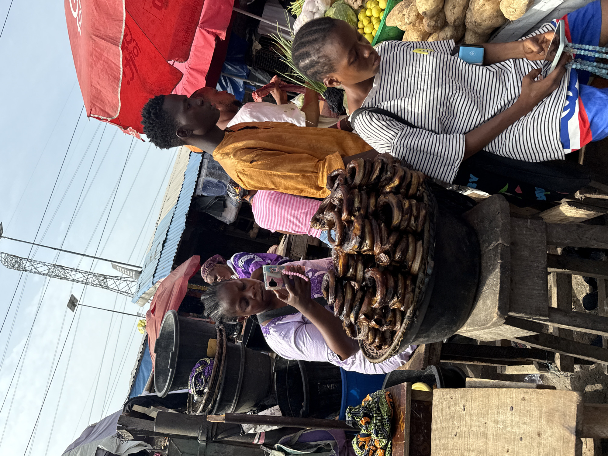 This image captures a vibrant outdoor market scene in Lekki, Nigeria, under bright daylight. The primary focus is on several individuals and various market goods.
In the foreground, a woman wearing a light purple dress with braided hair is looking at her smartphone, which has a pink beaded strap, possibly taking a picture or video. She is positioned next to a large black plastic tub. A large basket filled with numerous pieces of smoked fish, stacked high, is prominently displayed in the mid-ground.
To the right, another person, wearing a black and white striped shirt, is engrossed in their smartphone, holding it with both hands. Blue beaded bracelets are visible on their wrist, and they are seated on a blue and red patterned surface, with a small blue object next to them. Behind this individual, a person in a mustard-yellow shirt is also visible, looking towards the right.
The background reveals more of the bustling market environment, including stalls laden with produce such as light-colored tubers, green leafy vegetables, and yellow fruits. A large red umbrella, bearing a partially visible "Coca-Cola" logo, provides shade. Corrugated metal roofs and utility lines with a metal tower are also seen against a clear sky. The scene depicts typical daily life within an active market setting.