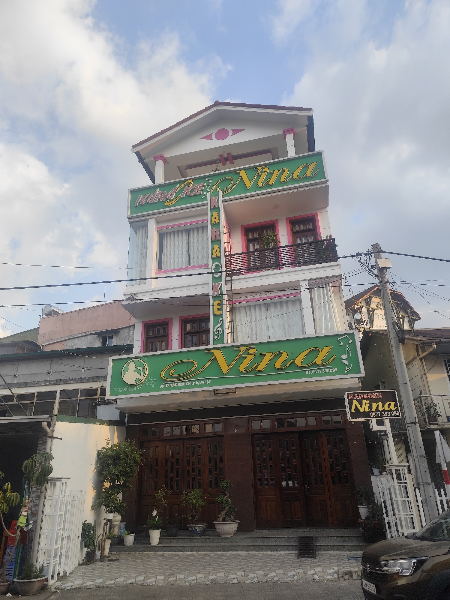 The image shows a three-story building with the name "Karaoke Nina" prominently displayed on green signs with gold lettering. The building has a white facade with pink accents around the windows and balconies. The address "17 Mac Dinh Chi, P.4, Da Lat" is visible on one of the signs, placing it in Da Lat, Vietnam. A vertical sign spells out "Karaoke" with a musical note. The entrance features dark wooden doors and potted plants. A smaller sign reading "Karaoke Nina" with a phone number is mounted on a utility pole. A car is parked in front of the building on the street. The sky is partly cloudy.