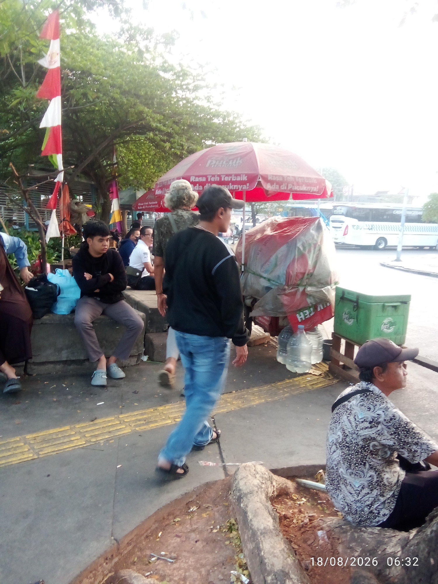 A group of people are gathered outdoors in what appears to be a market or roadside area. In the foreground, a man wearing a black shirt, blue jeans, and black sandals walks towards the right of the frame, away from the viewer. To his left, another man with dark hair and a black hoodie is seated on a concrete ledge with his arms crossed. He is wearing grey pants and grey Crocs. Behind them, several other individuals are visible, including a woman in a brown dress on the far left, and a person with grey hair wearing a camouflage-patterned shirt in the center. A red umbrella with text on it is partially visible above them. In the background, a white bus with green and black lettering is driving on a street. A green cooler box is positioned on the right side of the frame, next to a stack of items covered in red plastic sheeting. The ground is paved with concrete and yellow tactile paving. The scene is illuminated by bright, possibly late afternoon or early morning sunlight. The timestamp on the lower right corner indicates "18/08/2026 06:32". The location is identified as East Jakarta, Indonesia.