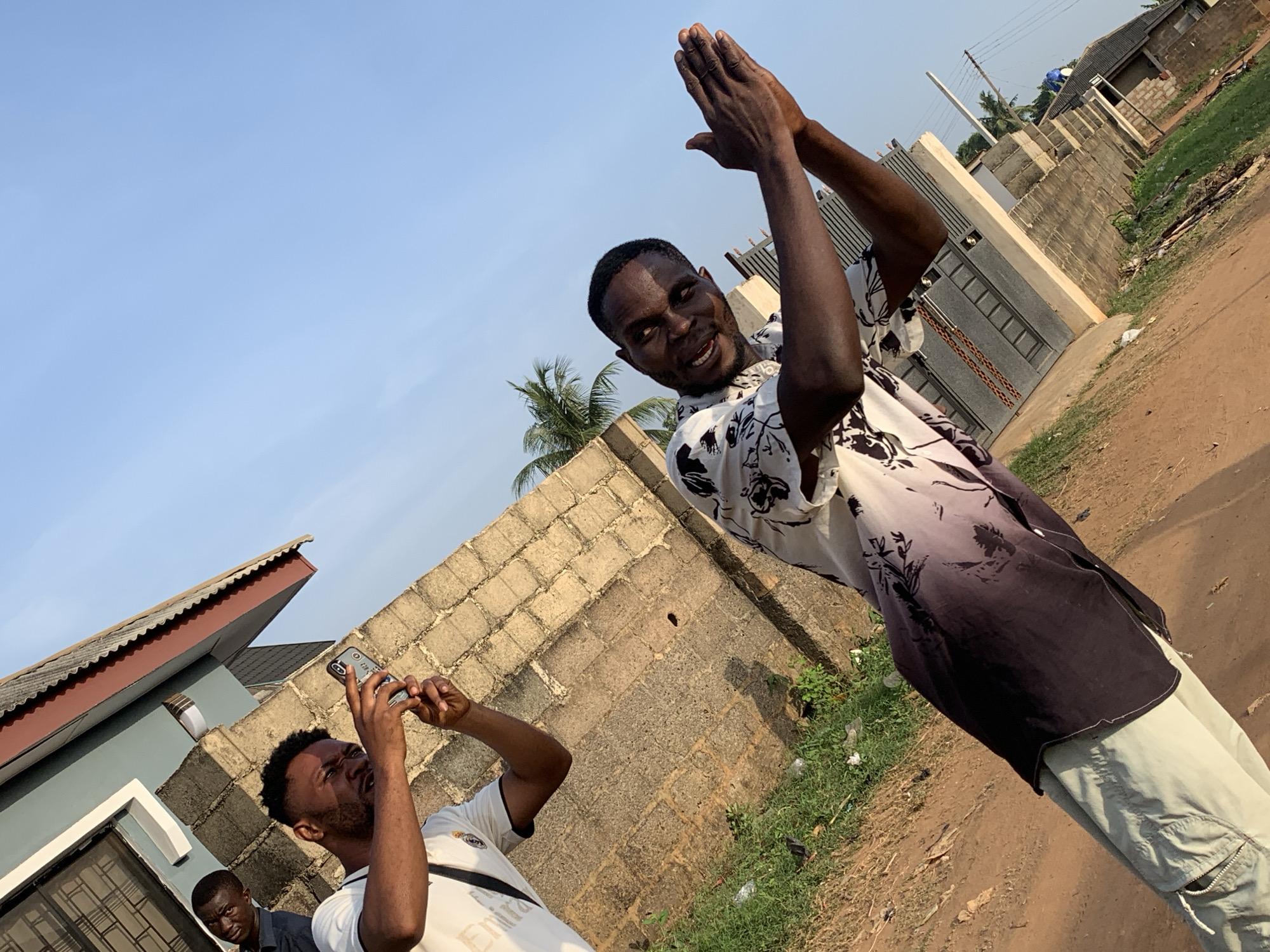 Three African men stand outdoors in Ikorodu, Nigeria. One man, in a white and black floral shirt and light pants, raises his hands up with the palms facing each other. A second man wearing a white shirt with writing on it holds up his smartphone to capture the moment. The third man, dressed in a dark suit coat, stands slightly behind the second man. A low brick wall and fencing are in the background. There is a low-rise building to the left. The ground is a dirt road with some green vegetation to the right. The sky is clear and light blue.