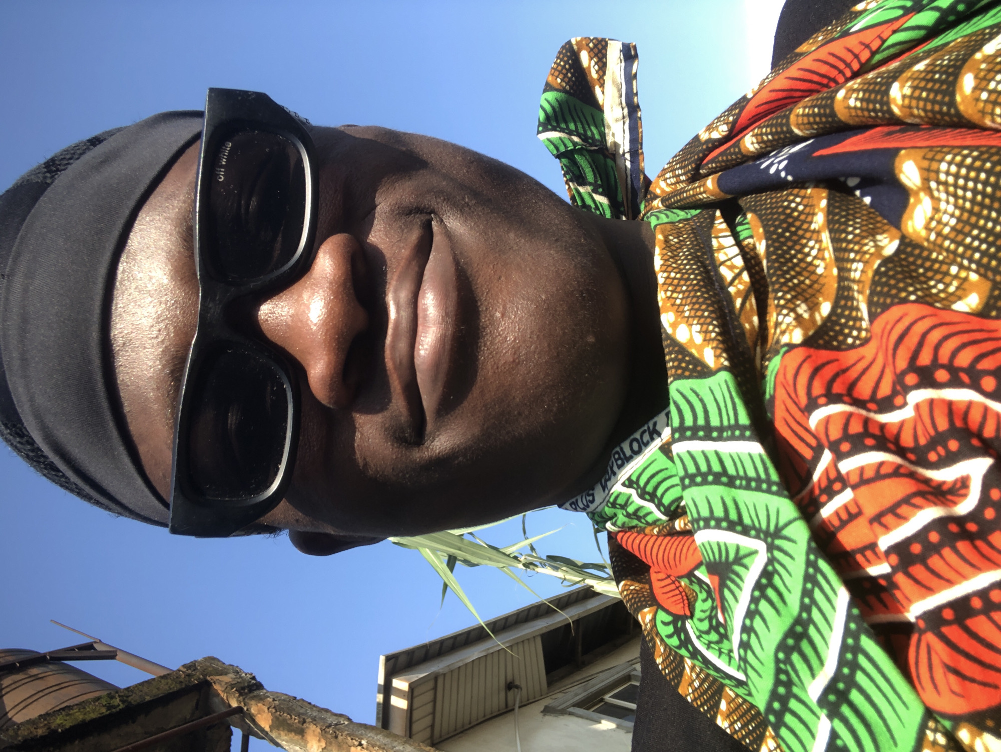 A close-up, selfie-style shot captures a Black individual wearing sunglasses and a black bandana. The individual is also wearing a brightly colored, patterned cloth wrapped around their neck, exhibiting vibrant green, orange, and yellow designs. In the background, a clear blue sky dominates, with a portion of a building and a water tank visible to the left, suggesting an outdoor urban setting, potentially in Ado, Nigeria, given the context. The sunlight illuminates the individual's face, creating highlights on their skin and reflecting off the sunglasses.