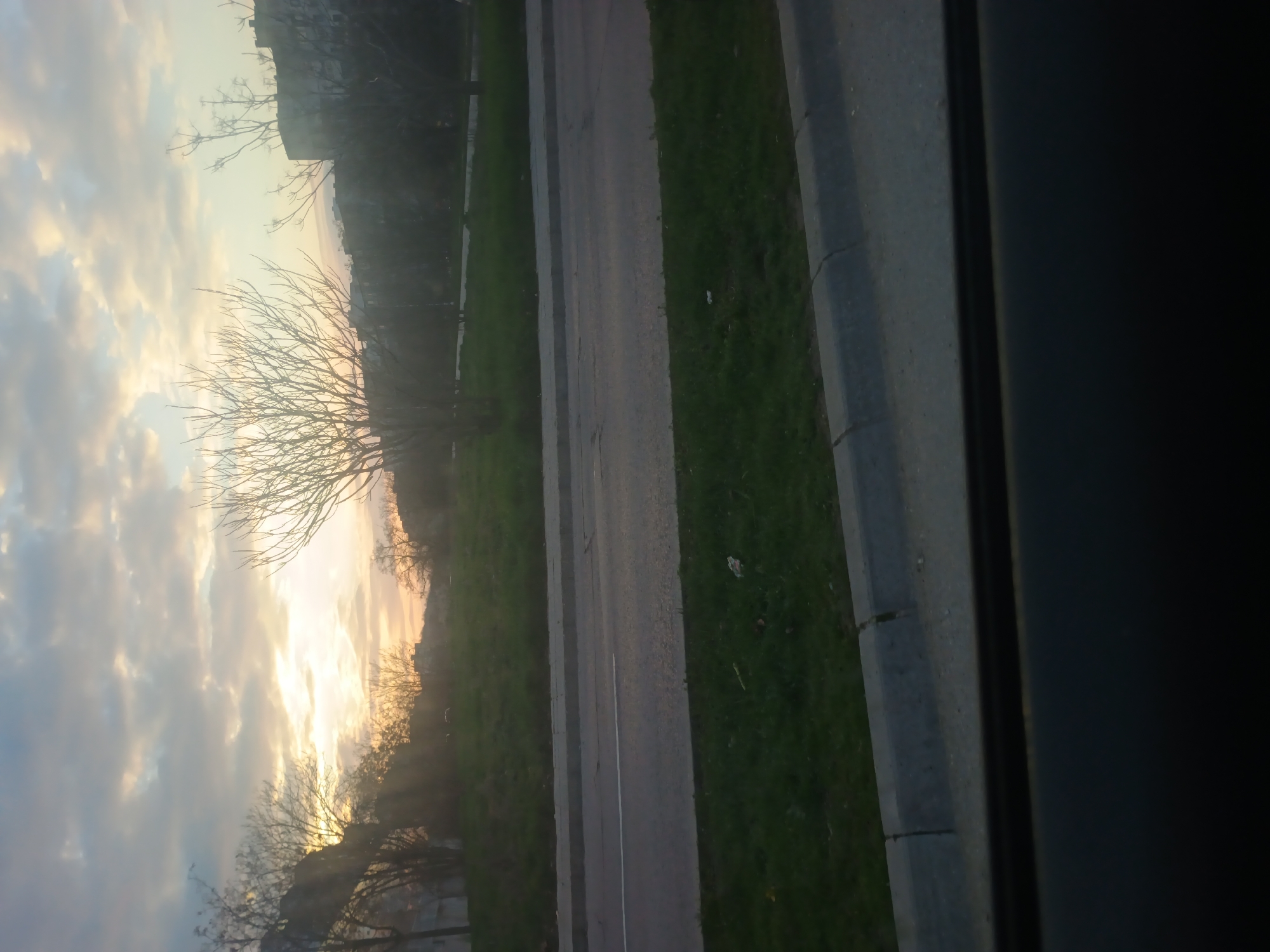 The image depicts a roadside view, likely taken from a moving vehicle on the outskirts of Varna, Bulgaria. The right side of the frame is occupied by the vehicle's window frame. To the left, a grey asphalt road runs parallel to a green verge. The sky is filled with scattered clouds during a sunset, casting a golden hue across the landscape. Bare-branched trees are silhouetted against the sky. Buildings are visible in the distance, suggesting an urban environment.