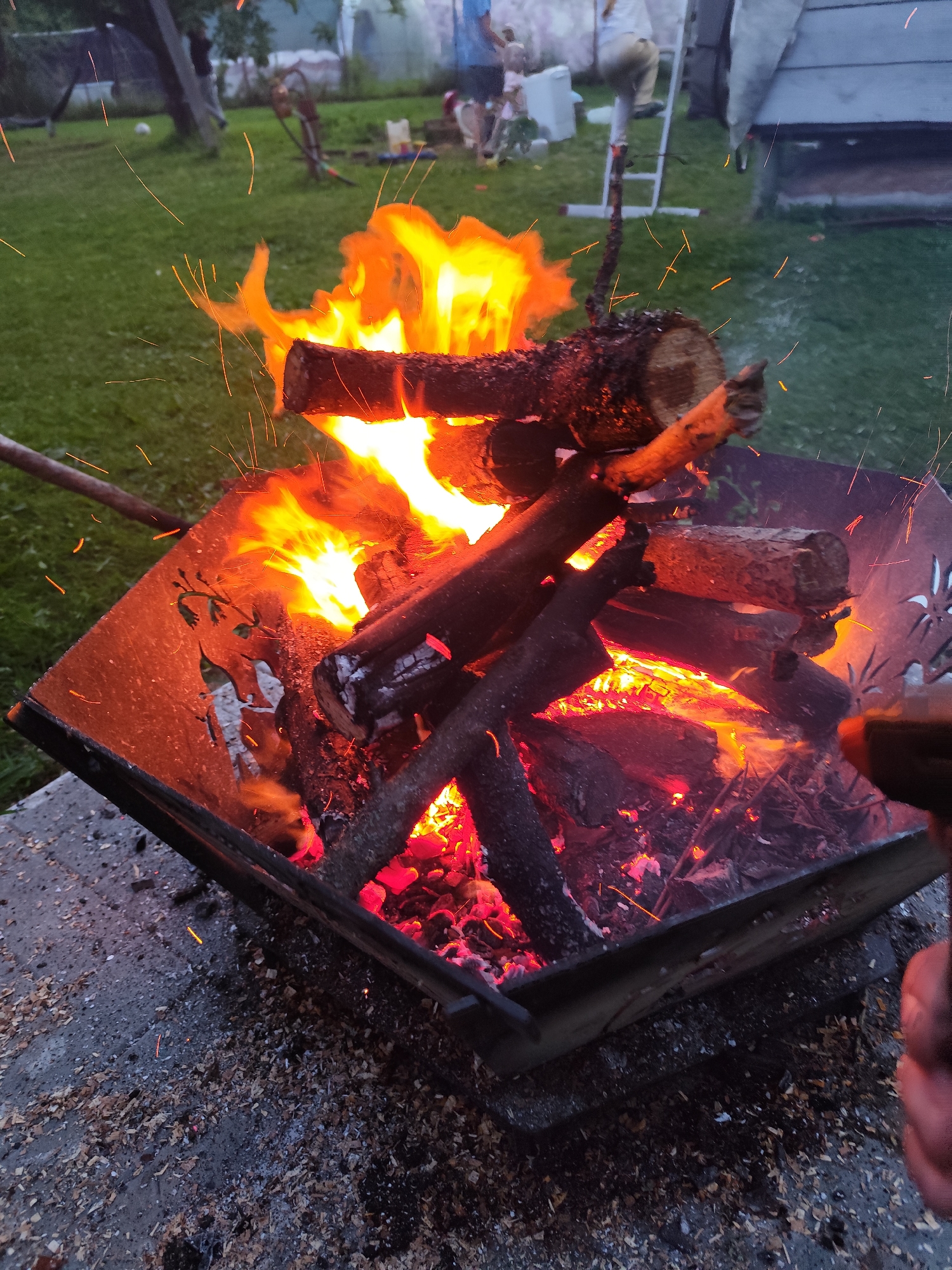 A fire pit is ablaze in a grassy backyard, with sparks flying upwards. The fire pit is square and made of metal, with a decorative pattern of leaves and figures cut into its sides. Logs are stacked inside the pit, burning intensely with bright orange and yellow flames. Ash and wood shavings are scattered on the concrete surface surrounding the fire pit. In the background, several people are visible, including a person standing on a ladder and others standing near a building and in the grass. The environment suggests an outdoor gathering or activity in a residential area. The specific location in Kolpakovo, Russia, indicates it is likely a dacha or a private home's backyard.