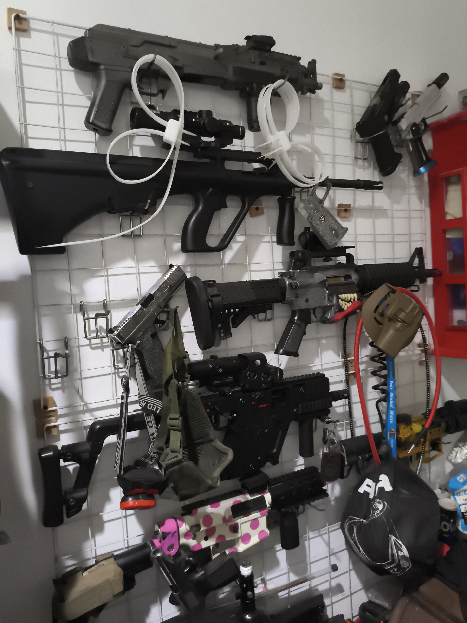 In the image, a wall-mounted display grid holds a collection of firearms, likely airsoft guns, arranged in rows. The grid is white and attached to a white wall with wooden pegs.
In the top row, a dark-colored rifle with a pistol grip is secured by white zip ties to the grid. Below it, a black rifle with a bullpup design is mounted.
In the middle row, a silver handgun is displayed, followed by a grey assault rifle with a black barrel and stock. A tan holster is attached to this rifle. To the right of the assault rifle, a red cabinet or structure is partially visible.
In the bottom row, another black rifle with a bullpup design is mounted. To its right, a smaller black firearm is visible, and further to the right, a black mask with white accents hangs from the grid. A gun accessory with pink and white polka dots is also visible.
Below these, parts of other firearms and accessories are present, including a tan stock and a black grip. The overall scene suggests a collection or display of replica firearms.
This image does not contain any identifiable landmarks or contextual clues that would allow for determining its location in West Jakarta, Indonesia.