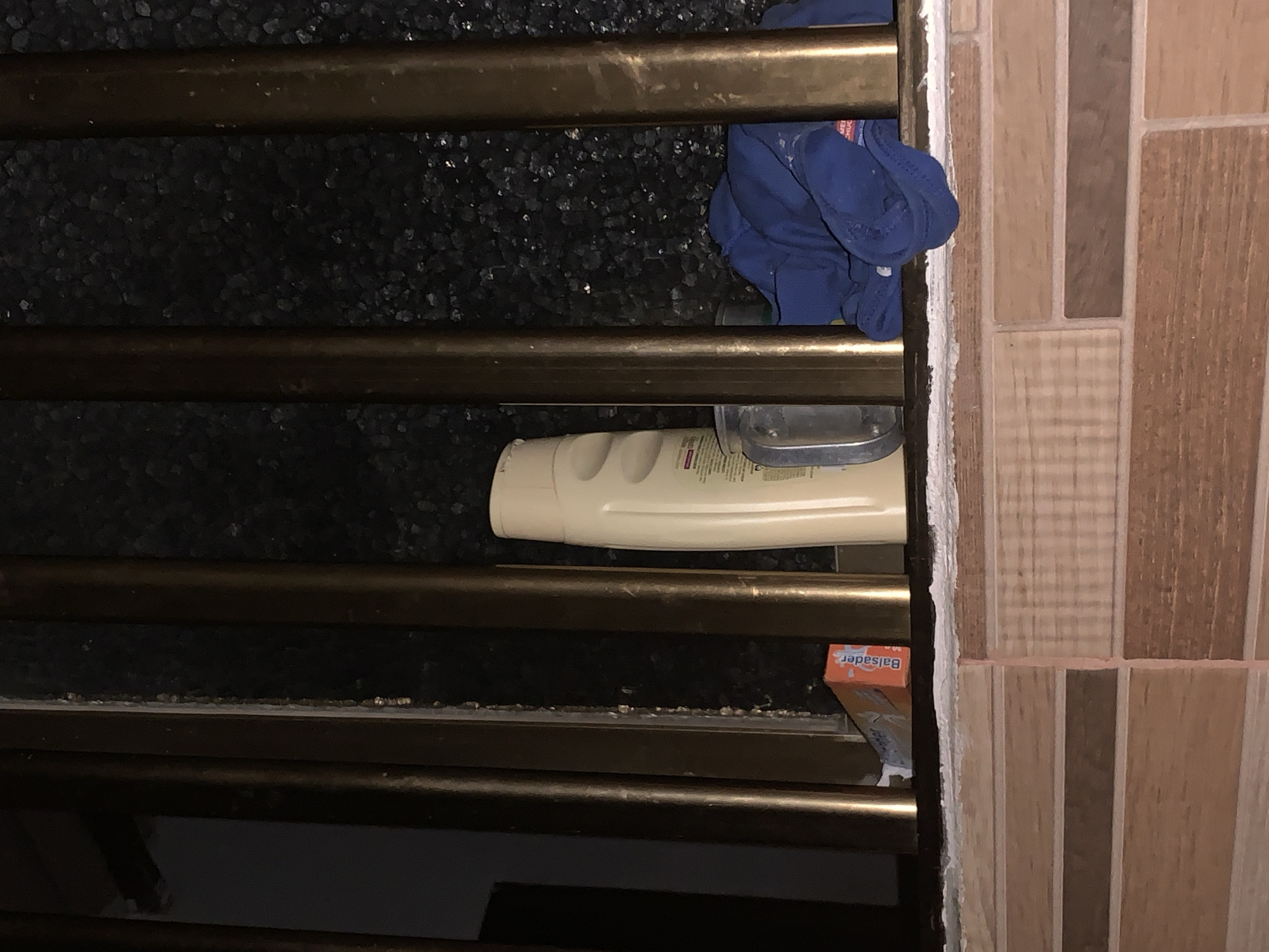 The image captures a static, close-up view of a section within what appears to be a bathroom or utility area, located in Santa Ana, Venezuela. In the foreground, several horizontal bronze-colored metal bars run across the frame, suggesting a towel rack, window grille, or part of a shower enclosure. These bars show signs of wear and possibly water marks.
Behind the bars, the background is a dark, textured surface, possibly a wall or flooring covered in small, dark, granular material that reflects some light. On the right side, a tiled wall is visible, featuring rectangular tiles in various shades of brown, beige, and tan, arranged in a staggered pattern with white grout lines. A thick line of white caulk or sealant is present where the metal bars meet the tiled wall.
Several objects are placed within this setting:
- A crumpled blue cloth or small towel is draped over one of the upper bars, on the right side.
- A large, off-white or light yellow bottle, likely shampoo or conditioner, rests horizontally on or behind one of the middle bars. Faint, illegible text is visible on its back label.
- Next to the white bottle, a silver-colored object, possibly a pump dispenser or another container, is partially obscured.
- A small orange box, with the word "Balsader" clearly printed in white letters, is positioned on a lower bar near the tiled wall.
No activity or event is taking place, and there are no people present. The lighting is ambient, not providing clues about the time of day or outdoor weather. The scene conveys a functional, everyday environment.