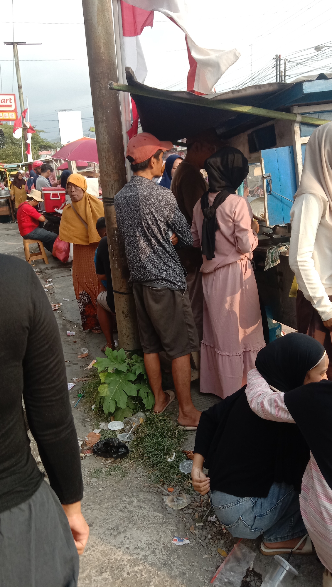 The image depicts an outdoor scene with several people gathered around a food stall. The ground is a mixture of concrete and gravel, with some patches of grass and debris. In the background, there are buildings and overhead wires, suggesting an urban or semi-urban environment.
Several individuals are visible. A person wearing a dark patterned shirt and shorts stands near a pole. To their right, a person in a pink dress and a black headscarf is interacting with someone at the stall. A person in a yellow headscarf and orange skirt is also present. Further left, a person in a red shirt and hat is seated. In the foreground, a person in a black long-sleeved shirt and grey pants is partially visible, and to the right, another person in a black headscarf and jeans is crouched down, leaning on someone wearing a striped shirt.
The food stall appears to be simple, with a dark covering and a blue structure behind it. There are various items on and around the stall, including what look like containers and possibly food items. Indonesian flags, red and white, are visible in the background, suggesting a patriotic or festive occasion. The overall impression is of a bustling marketplace or street food vendor area.