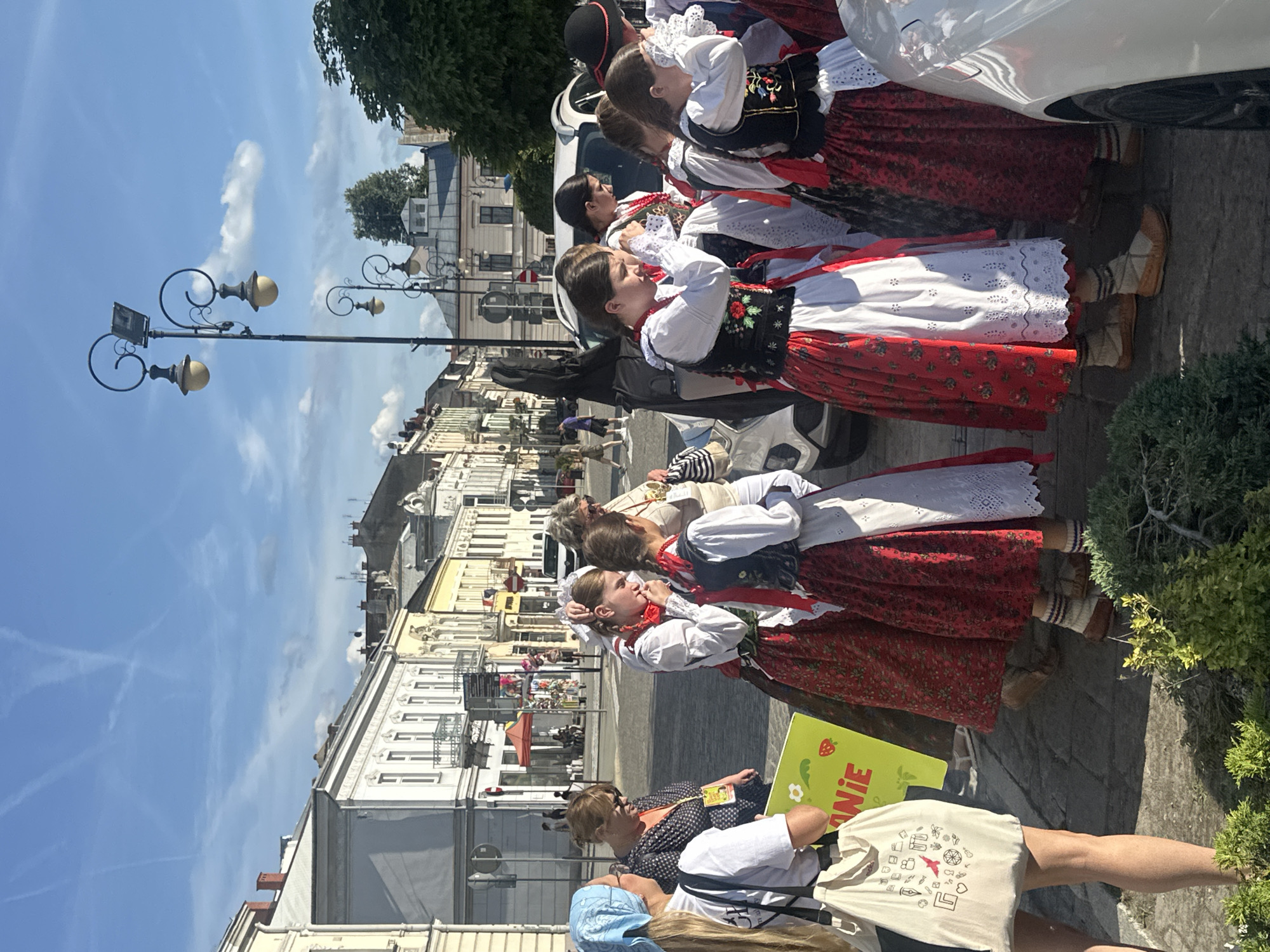 In Nowy Sącz, Poland, a group of young women dressed in traditional folk costumes stand on a street. The costumes feature red skirts with floral patterns, white blouses, and black vests adorned with embroidery. Some of the women have red ribbons in their braided hair. They are standing near parked cars and a white SUV. In the background, there are buildings with ornate architecture, streetlights, and a blue sky with some clouds. One woman on the left is wearing a light blue headscarf and a white t-shirt, carrying a tote bag with various drawings. Another woman is wearing sunglasses and a dark patterned shirt. The scene appears to be a public gathering or event in the city center, possibly a cultural festival or parade.
