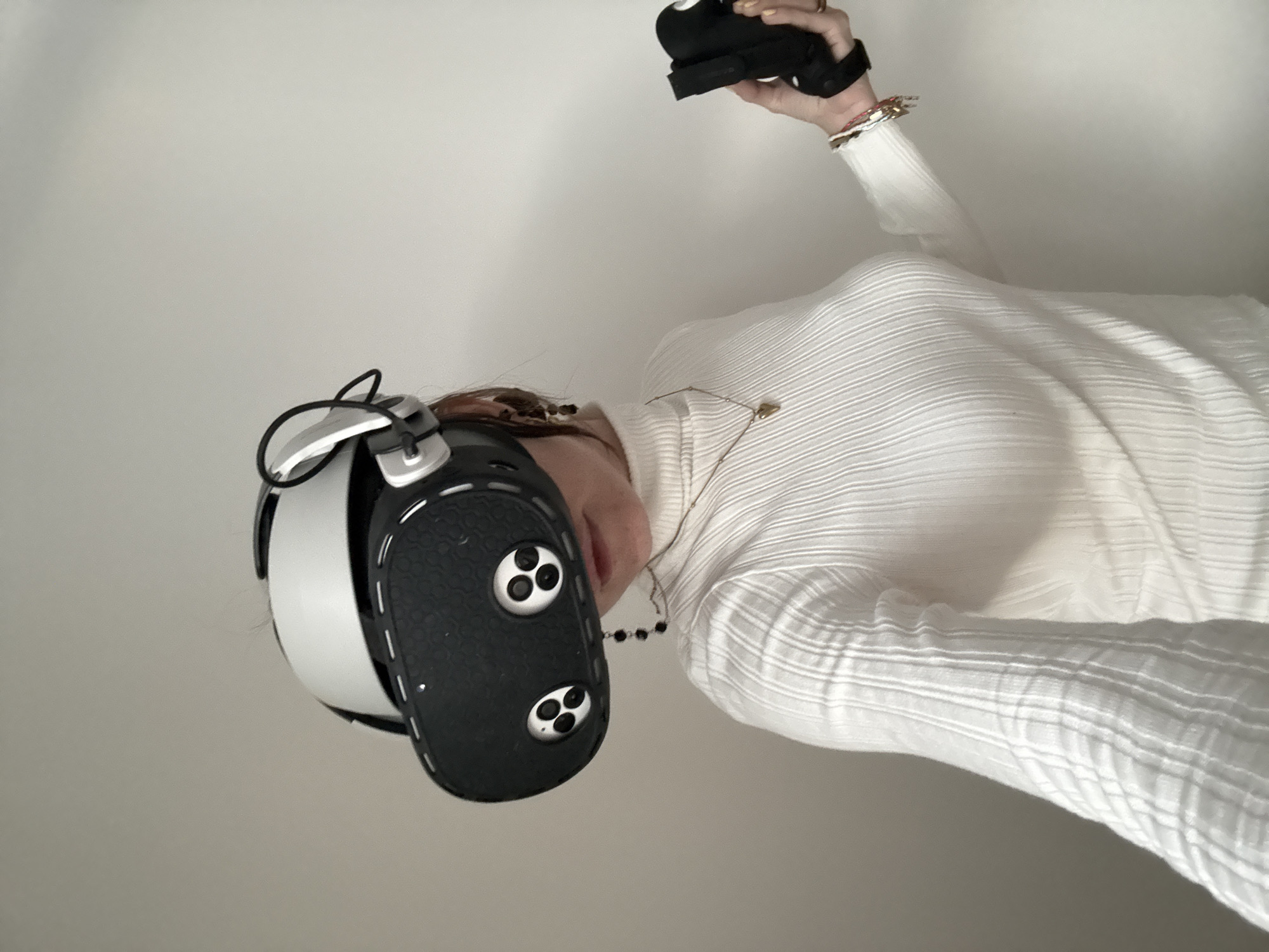 A person in a white turtleneck sweater is wearing a VR headset and holding a VR controller in their right hand. The headset is black and white with a textured surface. The controller is black with a strap around the hand. The person is facing left of the frame. The background is a plain, light-colored wall. The setting could not be identified as Moscow, Russia, due to the lack of identifiable landmarks or characteristics.