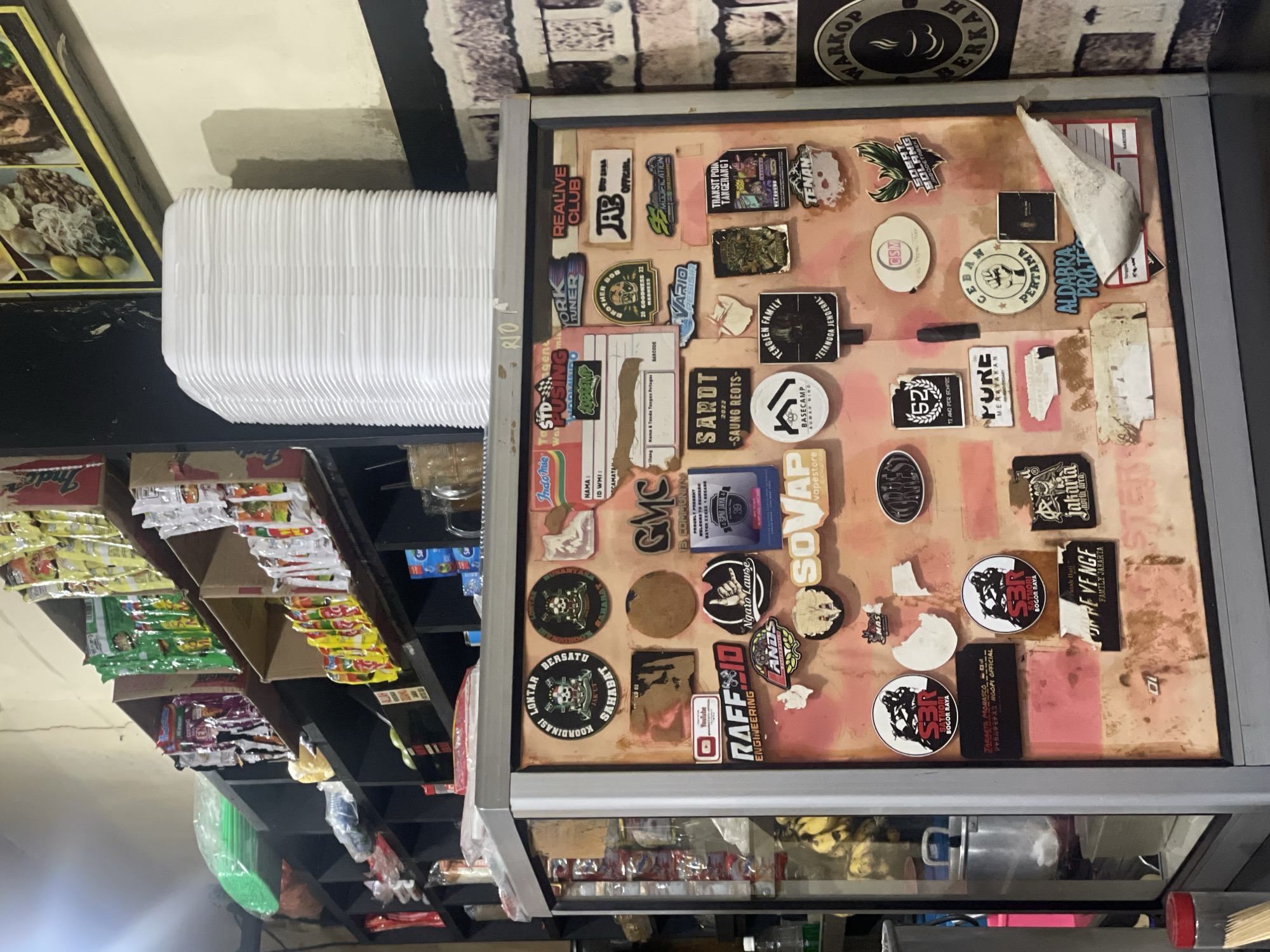 The image depicts a store interior, likely in North Jakarta, Indonesia based on local brand stickers and food items. A display case with a collage of stickers, possibly collected by the shop owner over time, dominates the center of the frame. The stickers range from local businesses, clubs, and slogans, to recognizable brands. Shelves stocked with snacks, instant noodles (Indomie brand), and drinks are visible to the left. A stack of white containers sits above the shelves. The environment suggests a small, local business, perhaps a convenience store or cafe. The scene conveys a sense of Indonesian local culture and commerce.