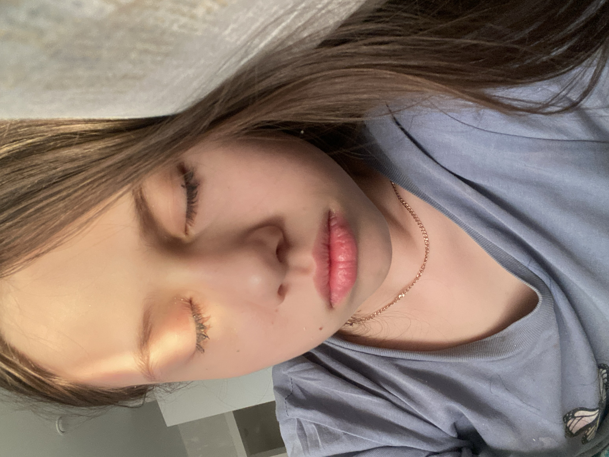 A close-up, sideways portrait shows a young person, likely a child or pre-teen, sleeping or resting indoors. Their eyes are gently closed, conveying a peaceful expression. The individual has long, straight, light brown hair that cascades over their shoulders and onto a light-colored surface, possibly a bed or pillow. They are wearing a light blue or grey t-shirt, with a partial, faded butterfly graphic visible on the fabric towards the bottom right of the frame. A delicate, thin gold-toned chain necklace is worn around their neck. Soft, warm light illuminates the left side of their face. The background is indistinct, suggesting an indoor environment. The scene is located in Izhevsk, Russia. No specific time of day or weather conditions can be determined from the image, and there is no visible text.