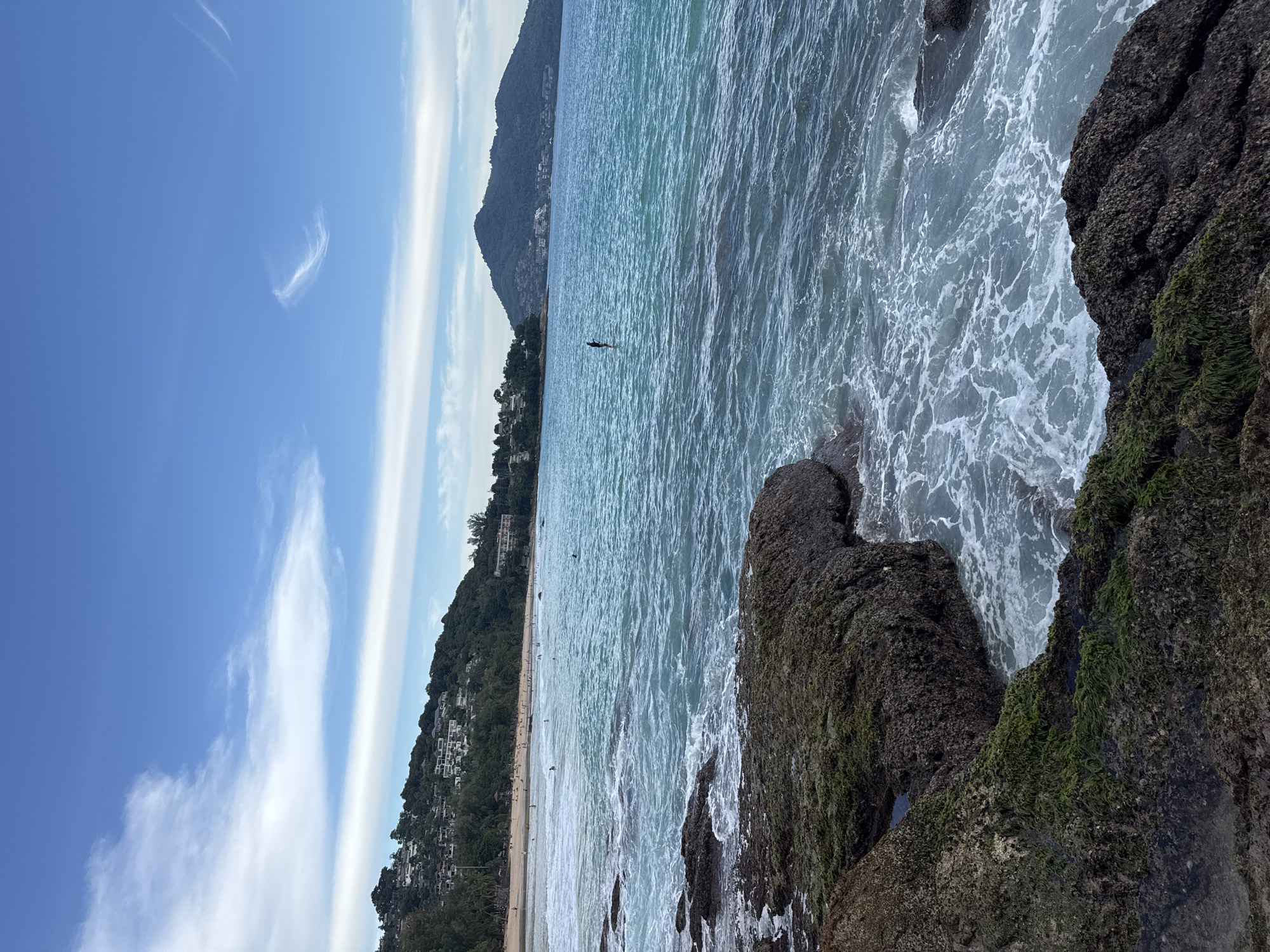 The image depicts a coastal scene at a beach in Choeng Thale, Thailand. In the foreground, dark, moss-covered rocks are partially submerged in the turquoise water, with white foam from the waves washing over them. Further out, the ocean is a vibrant blue, with gentle waves. A single person is visible in the distance, standing on a paddleboard. Beyond the water, a sandy beach stretches along the coastline, dotted with numerous people. Lush green hills, covered in trees and buildings, rise behind the beach, suggesting a developed resort area. The sky above is a clear blue with scattered wispy white clouds. In the far distance, a large, tree-covered mountain dominates the horizon.