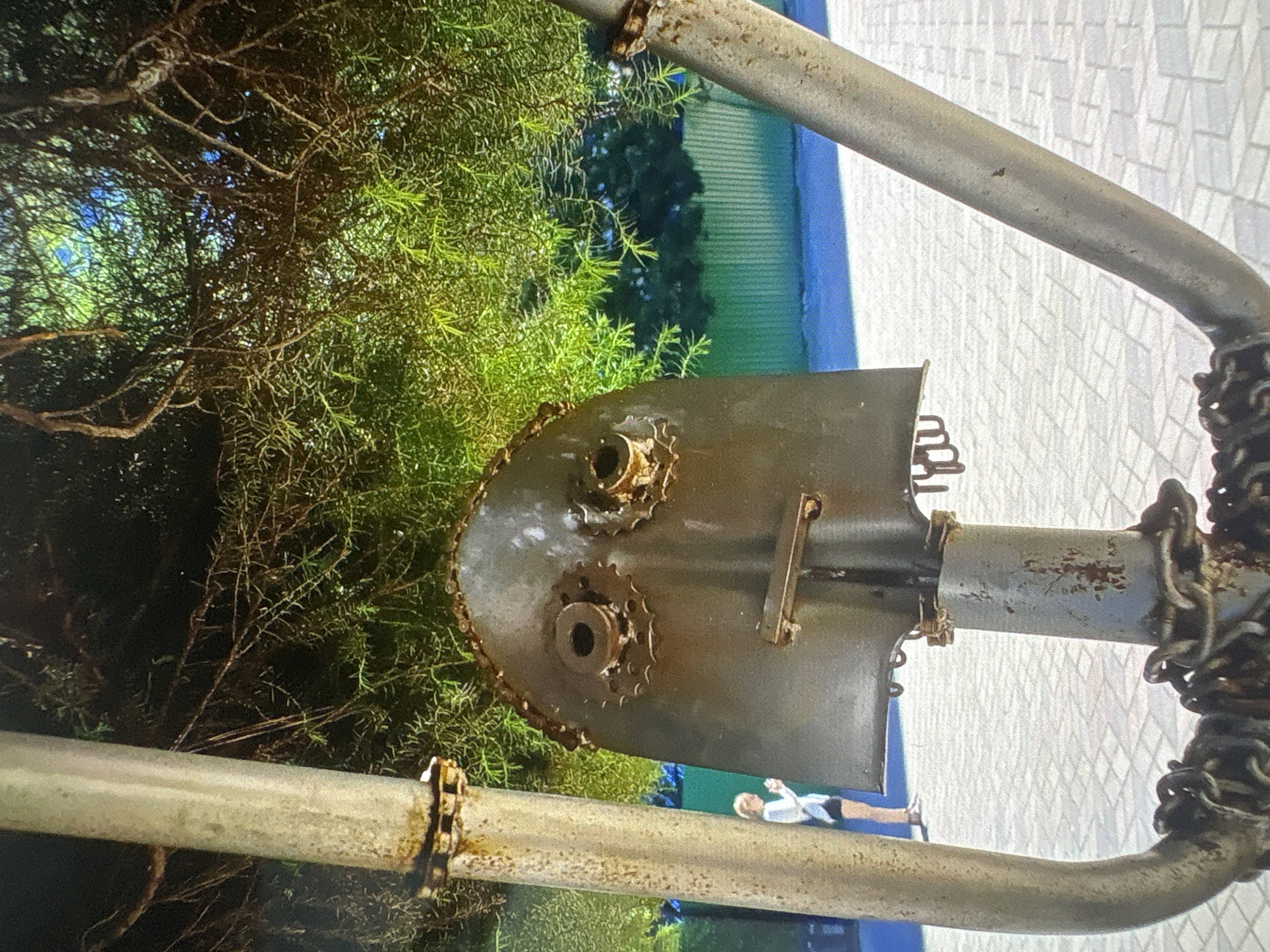The image shows a close-up of a metal sculpture resembling a face, constructed from what appears to be a shovel head for the face, gears for eyes, and a bolt for a nose. The sculpture is outdoors, with green foliage from a tree overhead. In the background, a young person wearing a white top and dark shorts stands on a paved walkway. A green corrugated fence is visible behind the walkway. The paving stones are light-colored and arranged in a grid pattern. The sculpture is metallic and shows signs of rust. The overall scene suggests an outdoor public space.