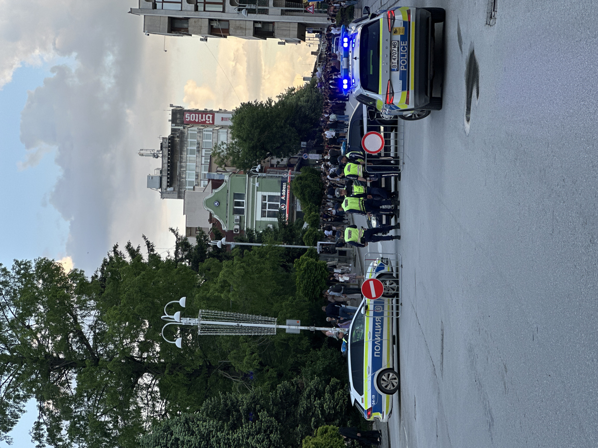 The image shows a street in Veliko Tarnovo, Bulgaria, where a crowd of people is gathered. Two police cars are parked on the street, with police officers standing nearby, appearing to form a barrier. The first car has a "no entry" sign attached to the barrier next to it. The second car has its emergency lights on. Buildings and trees line the street in the background. "Britos" is written on one of the buildings in the background. The sky is cloudy.