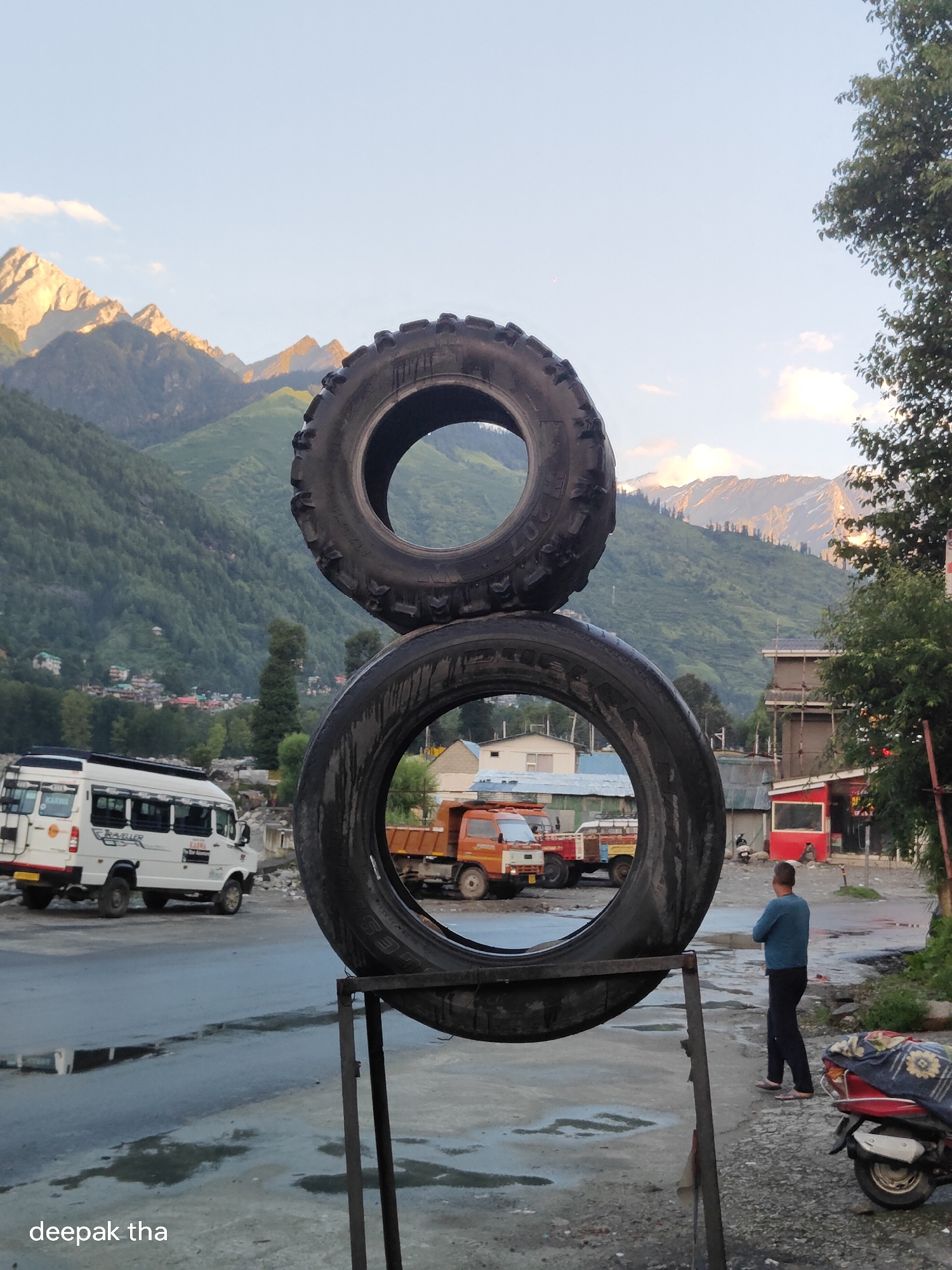 In Vashisht, India, two large tires are stacked vertically, one on top of the other, forming an infinity symbol. The tires are positioned in the foreground, obscuring parts of the background. Behind the tires, a road is visible, wet with puddles reflecting the sky. A white van is parked on the left side of the road. Further back, several trucks are parked, and buildings with corrugated roofs line the street. In the distance, a man in a blue shirt and dark pants stands facing away from the camera, looking towards the buildings. A red scooter is parked to his right. The background is dominated by lush green mountains with snow-capped peaks under a clear sky with scattered clouds. The lighting suggests it is late afternoon or early evening.