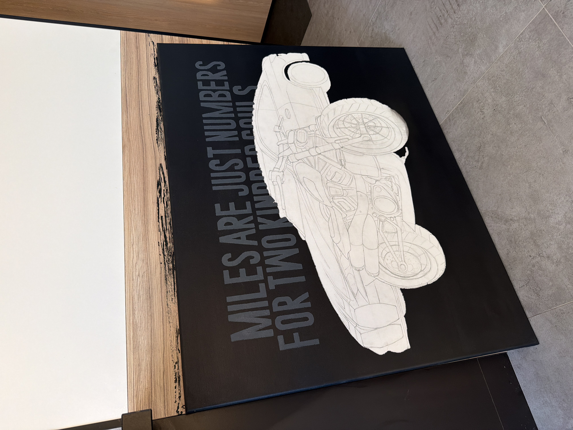 The image shows a black canvas with a white sketch of a motorcycle and the text "MILES ARE JUST NUMBERS FOR TWO KINDRED SOULS". The canvas rests on a table next to a white and wood-grain surface. The floor is tiled with gray tiles. The location is Unknown, Unknown.