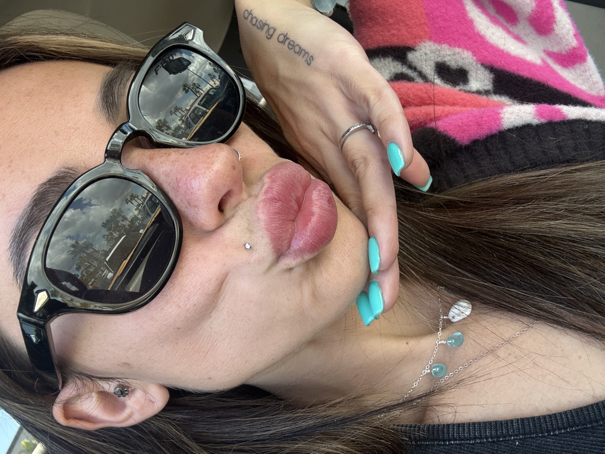 A person, possibly female, is posing in an image. The person is wearing sunglasses that reflect an exterior scene of palm trees, parked cars, and what seems to be buildings under a sunny sky, which is indicative of the city of Long Beach, California, United States. She has pierced ears and a pierced lip. The person is also wearing a ring, and turquoise-colored nail polish on her fingernails. A tattoo that reads "chasing dreams" is visible on the person's right wrist. She is wearing a pink and dark-colored top. There is also a delicate silver necklace around the neck.