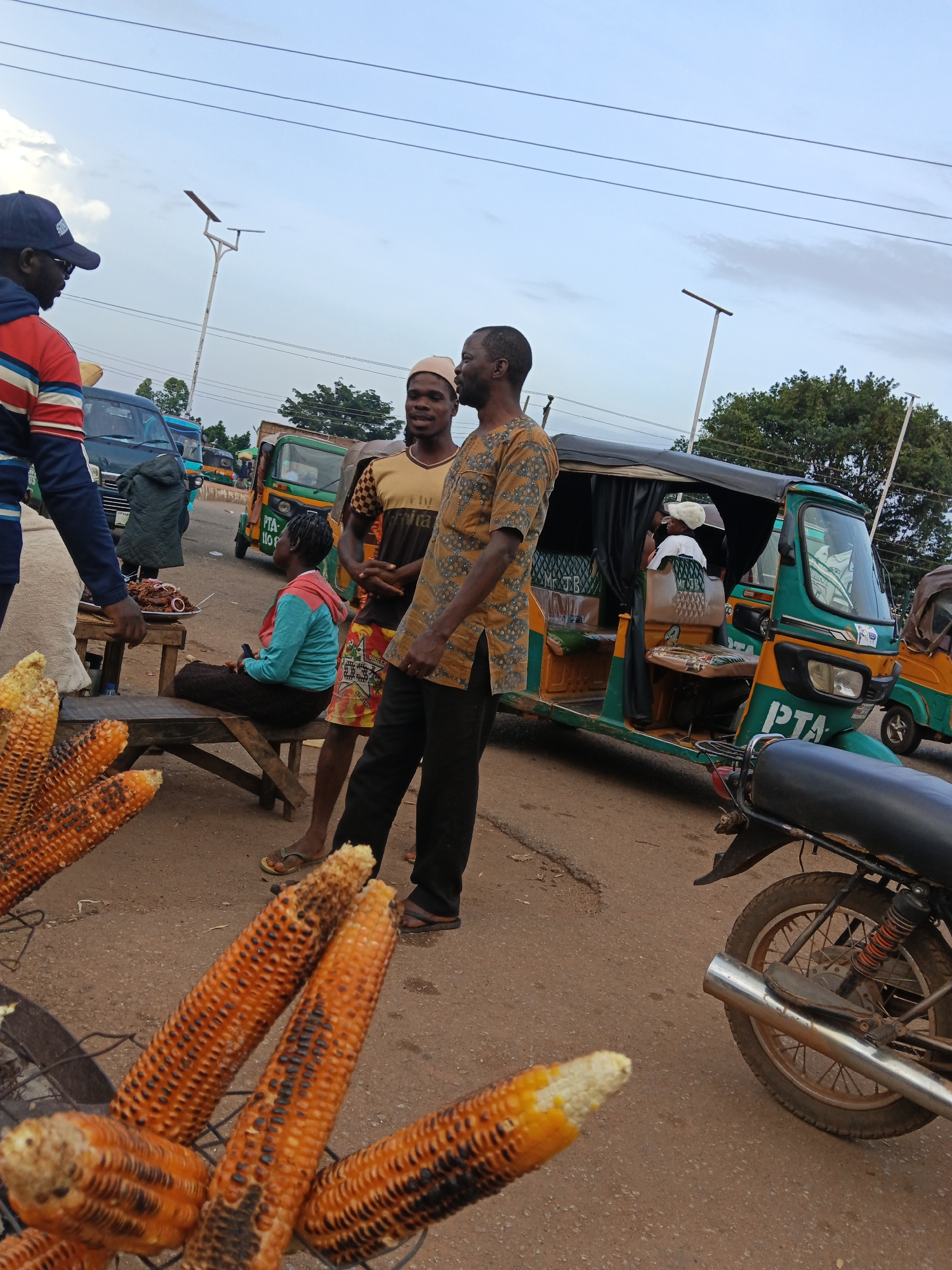 At an outdoor market in Bukuru, Nigeria, a group of people are gathered around a street food stall. In the foreground, several ears of roasted corn are displayed on a wire rack. To the left, a man wearing a blue and red striped jacket and a baseball cap stands with his back to the camera. He is looking towards a table displaying food. Seated on a wooden bench is a woman in a blue hooded jacket and a dark skirt, with her hair covered. A man in a patterned brown shirt is standing next to her, looking towards the right. Behind them, two men in traditional attire are talking. A green and yellow auto-rickshaw, a common form of transport in Nigeria, is parked nearby. Several other vehicles, including a blue bus, are visible in the background, suggesting a busy thoroughfare. The sky is overcast, and the ground is paved with reddish-brown earth. The overall scene depicts a typical street market environment with vendors and customers interacting.