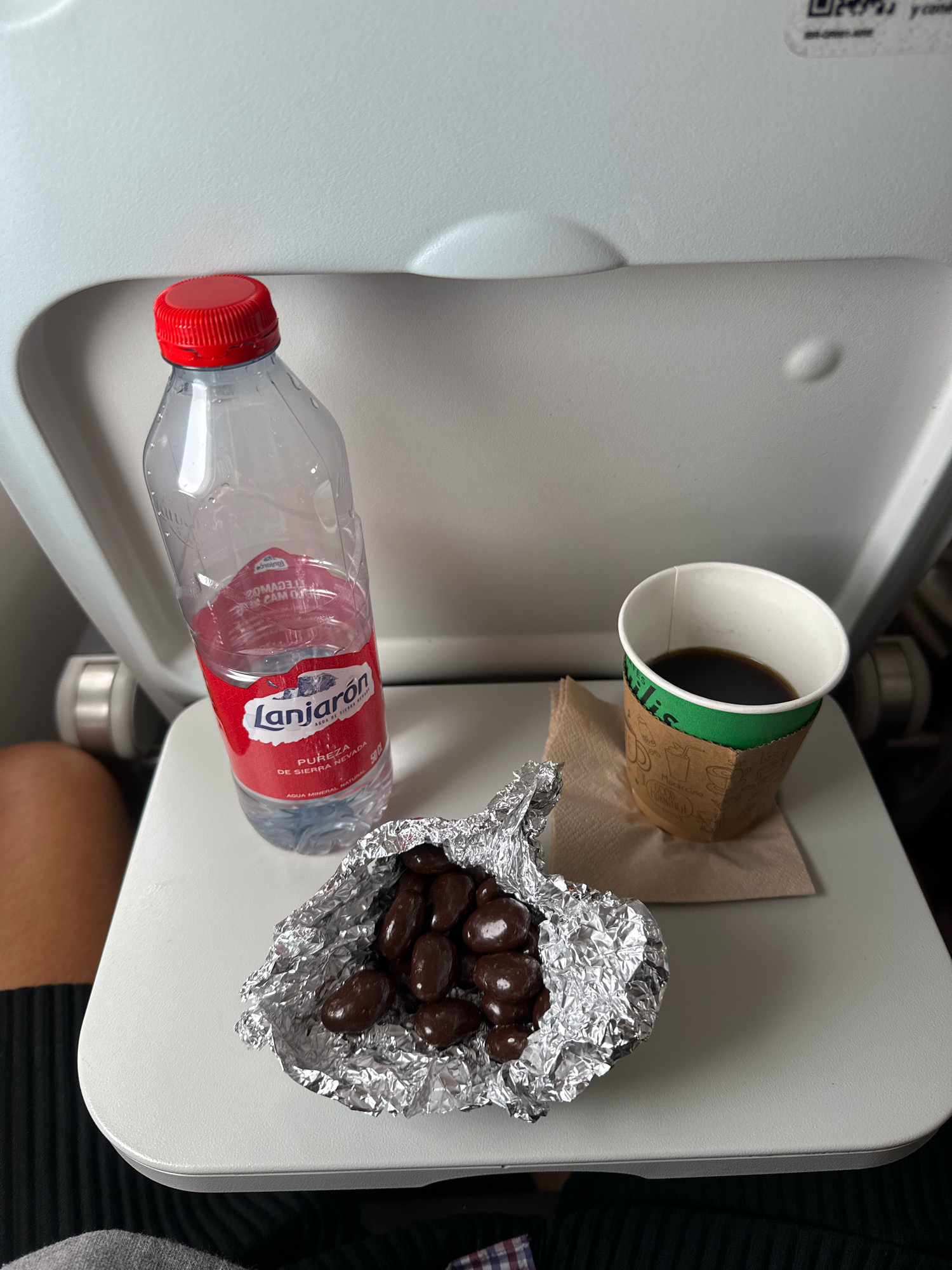 The image shows a tray table on an airplane with a bottle of Lanjarón water, a cup of black coffee on a napkin, and a small packet of chocolate-covered nuts wrapped in aluminum foil. The tray table is white and appears to be made of plastic. The background is a gray airplane seat. There is no indication of the city of Ciudalcampo, Spain in the image.