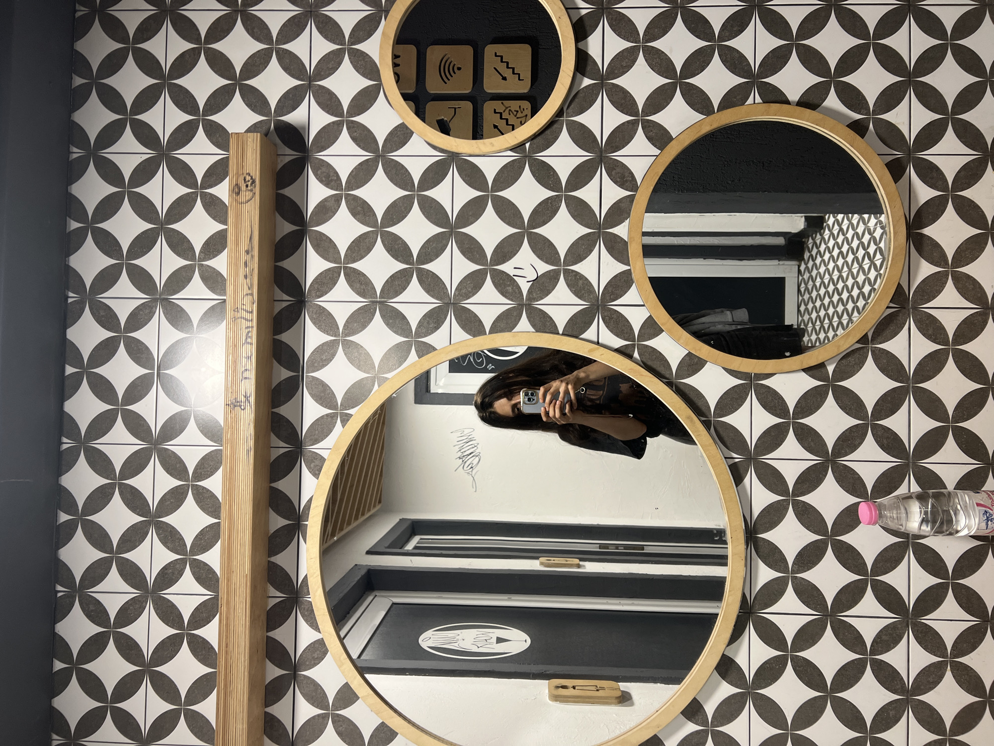 The image depicts a woman taking a selfie in a restroom with distinctive black and white geometric patterned tiles on the wall. The woman is holding a smartphone in her right hand and appears to be wearing a dark t-shirt. In the foreground, on the bottom left, is a clear plastic bottle of water with a pink cap. There are three circular mirrors with wooden frames mounted on the tiled wall. The largest mirror, in the center-left, reflects the woman and two doors behind her, one of which has a circular logo with a wine glass and a silhouette of a person. A wooden shelf is mounted above the largest mirror, with some graffiti on it. To the right of the large mirror, there are two smaller square wooden plaques with symbols, including Wi-Fi and stairs, and a third, smaller circular mirror. A faint smiley face is drawn on the tiles between the large mirror and the smaller square plaques. The floor, visible in reflections, is tiled with a similar geometric pattern. The overall impression is of a modern, stylish restroom environment. This scene is located in Sofia, Bulgaria.