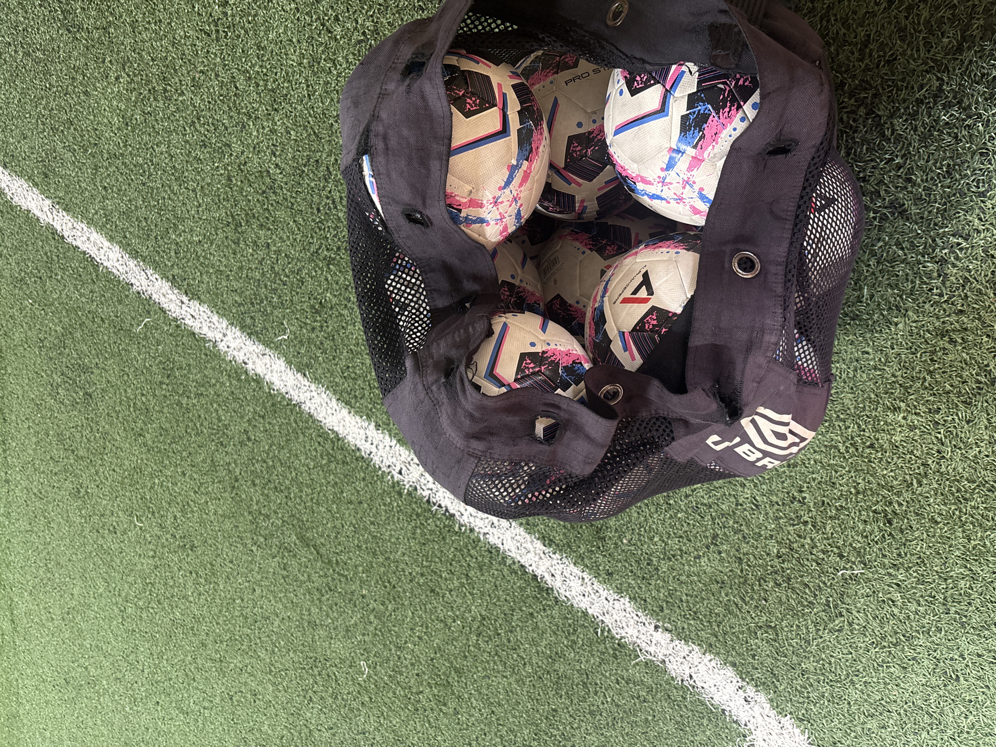 A mesh bag filled with several soccer balls lies on a green artificial turf field marked with a white line. The soccer balls have a white, pink, and blue design. The bag is black and has some lettering on it that is partially obscured. The scene appears to be an outdoor sports facility. There is no indication of the location being in Chaikovsky, Russia.