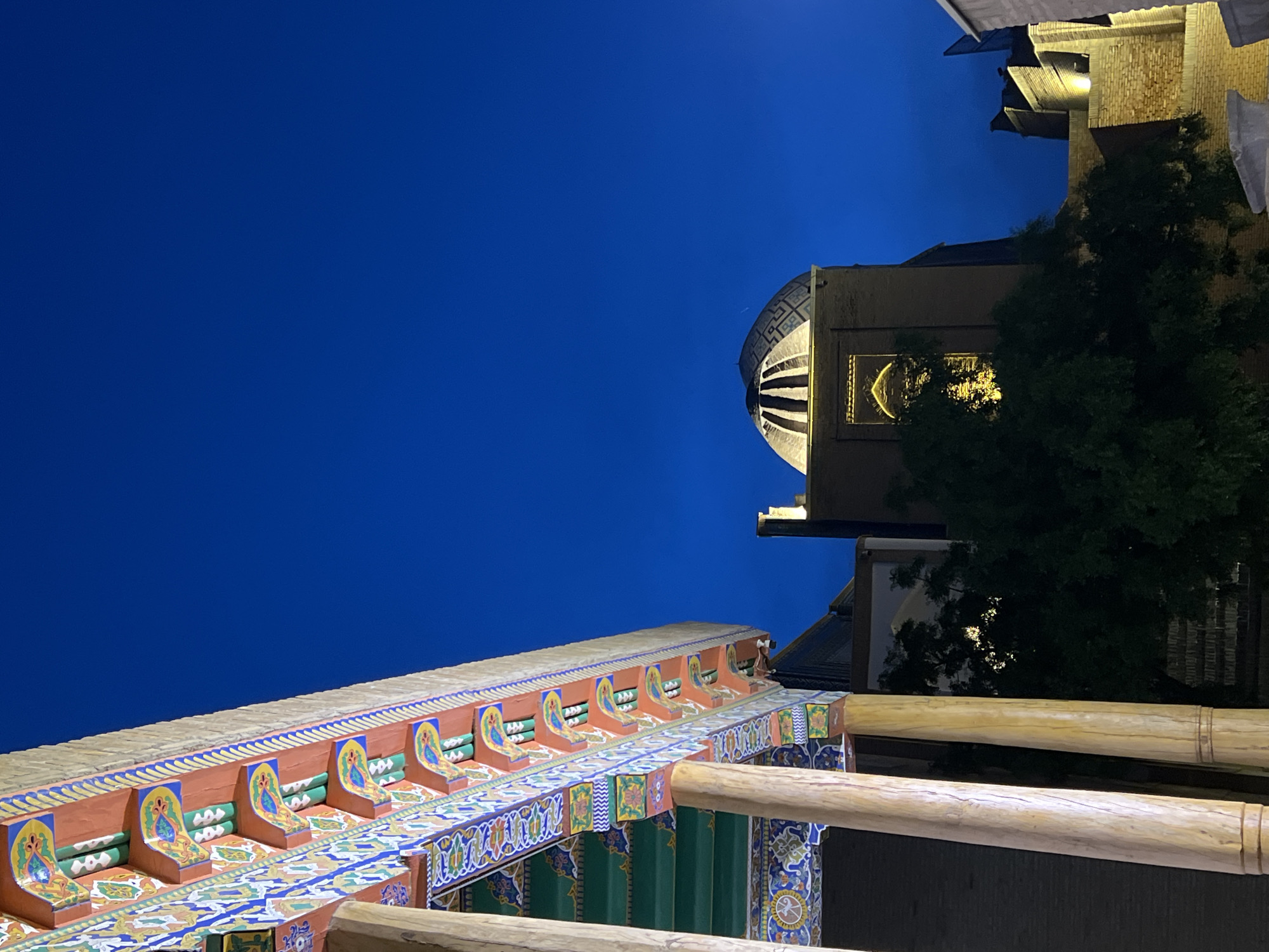 The image depicts the ornate architecture of Samarkand, Uzbekistan, at dusk. The vibrant blue sky dominates the upper portion of the frame, suggesting a clear evening. In the foreground, a brightly colored, intricately decorated wooden structure extends across the lower half of the image. This structure features detailed patterns in blue, yellow, green, and orange, with repeating motifs and what appear to be stylized figures. Wooden beams, possibly part of a balcony or roof, are visible in the lower right.
In the middle ground and background, the warm glow of artificial lights illuminates elements of a building, highlighting its brickwork and a large, domed structure with patterned detailing. A dark green tree stands to the right of this building, its foliage silhouetted against the illuminated architecture and the twilight sky. The overall scene evokes a sense of historical grandeur and cultural richness, likely a tourist site or religious building in Samarkand. There is no visible text in the image.