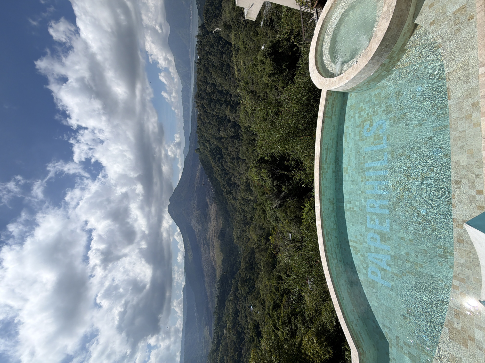 This is a photograph of a resort infinity pool and hot tub overlooking a lush green valley and mountains in Kintamani, Indonesia. The pool is filled with clear turquoise water and the word "PAPERHILLS" is visible on the pool floor in blue tiles. A small hot tub is attached to the side of the main pool. The background features dense green foliage covering rolling hills and a large volcanic mountain in the distance, with a lake visible beyond it. The sky is partly cloudy with scattered white and grey clouds against a bright blue sky. The image is taken from a high vantage point, suggesting a resort or hotel setting with a panoramic view.