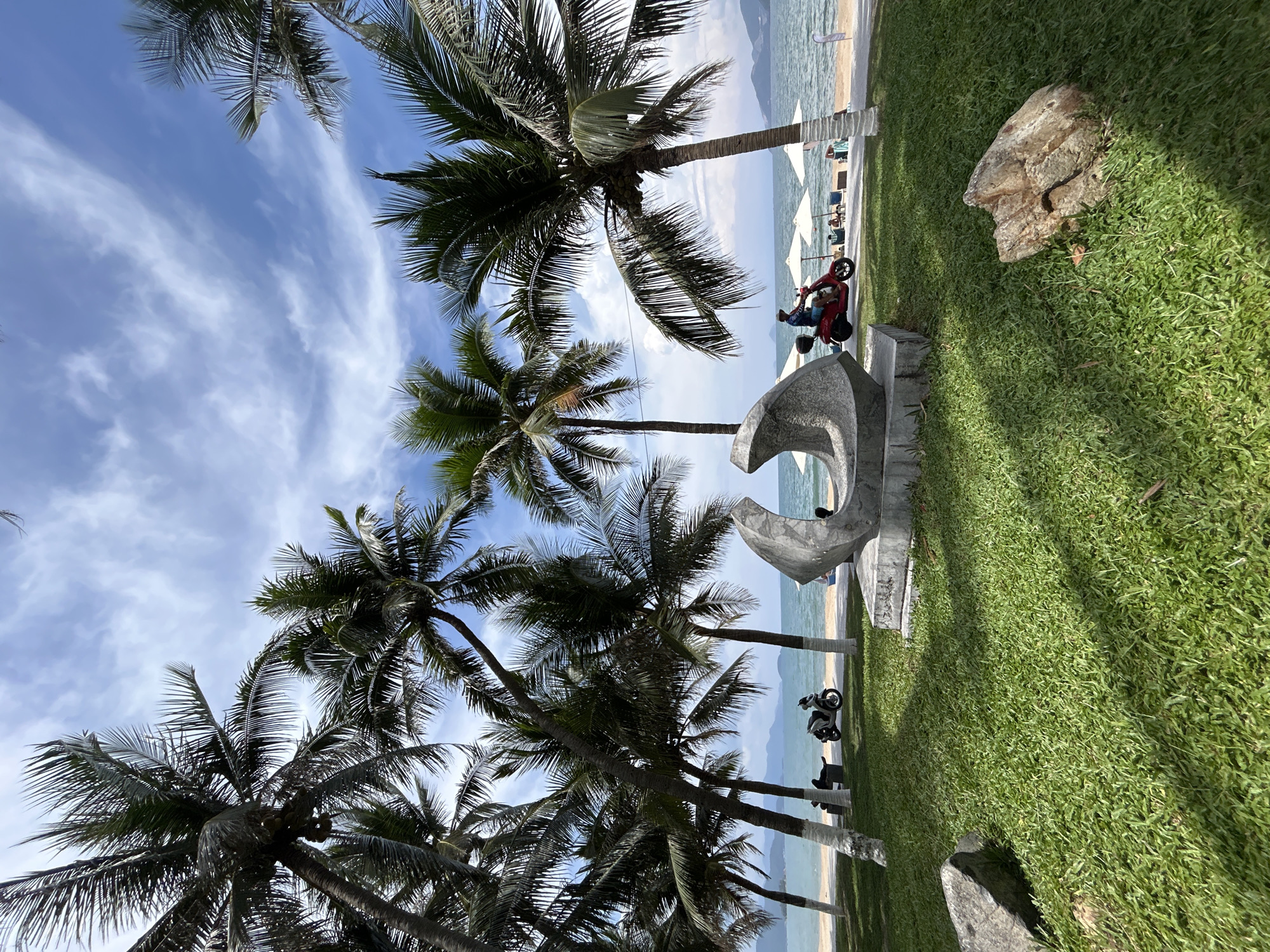 The image depicts a tropical scene, likely a resort area in Nha Trang Ward, Vietnam. Palm trees dominate the foreground, their silhouettes contrasting against a blue sky with wispy clouds. A sculpture, possibly made of concrete, sits on a grassy lawn. Further back, the beach is visible, with white umbrellas and lounge chairs suggesting a relaxation area. Two motorcycles are parked nearby. The photo seems to be taken from a low angle, looking upwards.