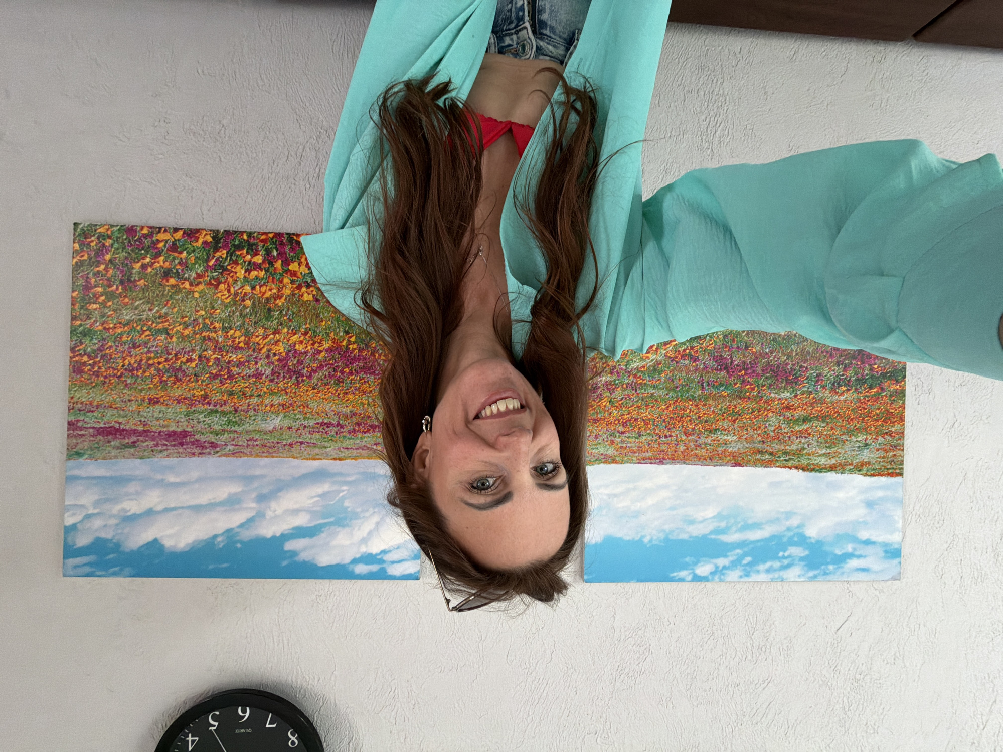 An outdoor, medium shot shows a Caucasian woman with long, brown hair and blue eyes taking a selfie in front of a wall with a large landscape picture. She is wearing a teal open-buttoned shirt over a red bikini top and jeans. Her left arm is extended towards the camera. A black analog clock hangs on the wall above the landscape picture. The overall lighting suggests daylight. The background shows a textured white wall and a framed image of a colorful flower field under a cloudy blue sky. The location is described as being in Birobidzhan, Russia, though no specific landmarks confirm this.
