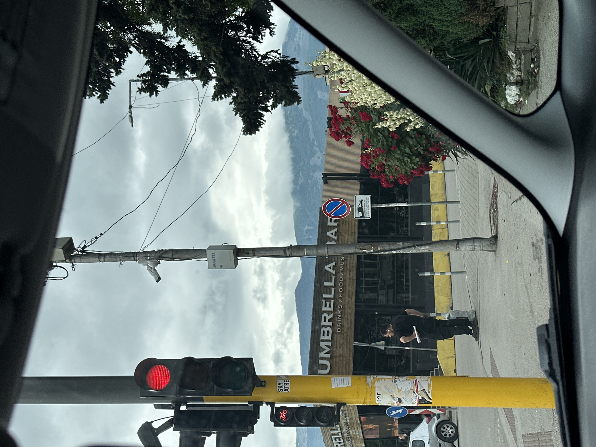 The image shows a street scene in Sofia, Bulgaria, on an overcast day. In the foreground, part of a car's interior is visible, including the windshield and dashboard. To the left, a yellow traffic light pole stands, with a red traffic light illuminated. A person in a black t-shirt and dark pants is standing on the sidewalk, facing away from the camera, with a piece of paper in their right hand. Behind the person is a building with the sign "UMBRELLA BAR DRINKS / FOOD / MUS". There are also several traffic signs and poles in the vicinity. In the background, a mountain range is visible, with clouds obscuring its upper parts. Trees and flowering bushes add to the urban landscape. The scene appears to be a typical street corner in a city, with a mix of infrastructure, commercial establishments, and pedestrian activity.