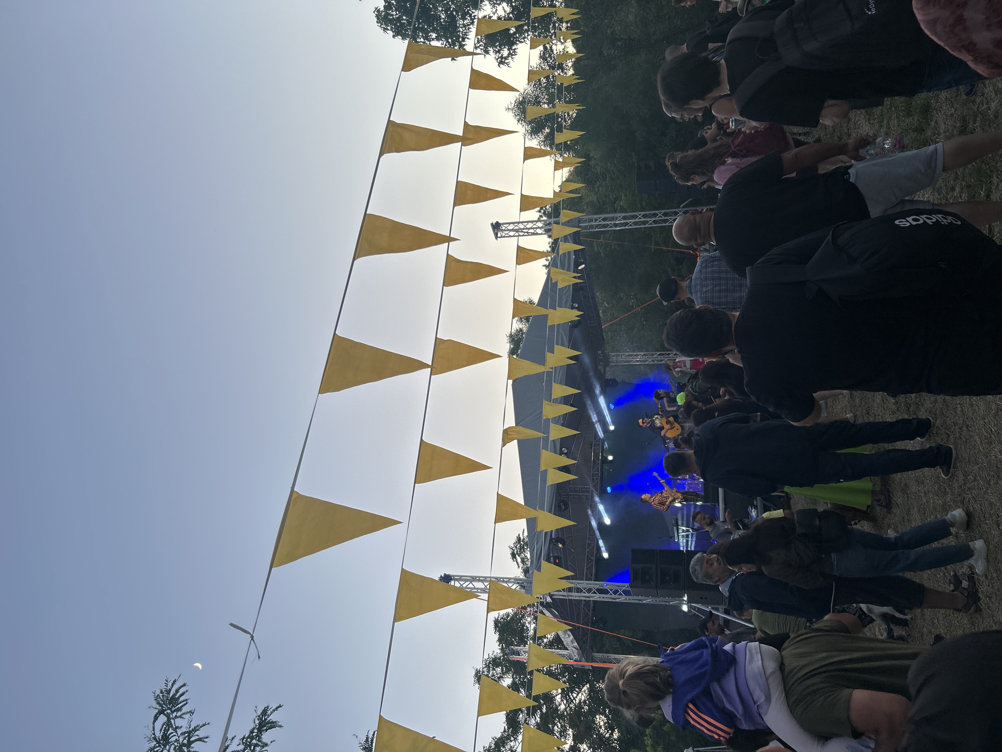 An outdoor concert event is underway with a band performing on a stage illuminated by blue lights. A crowd of people are gathered in front of the stage, watching the performance. Yellow triangular flags are strung across the sky, decorating the event space. The moon is visible in the upper left corner of the image.
